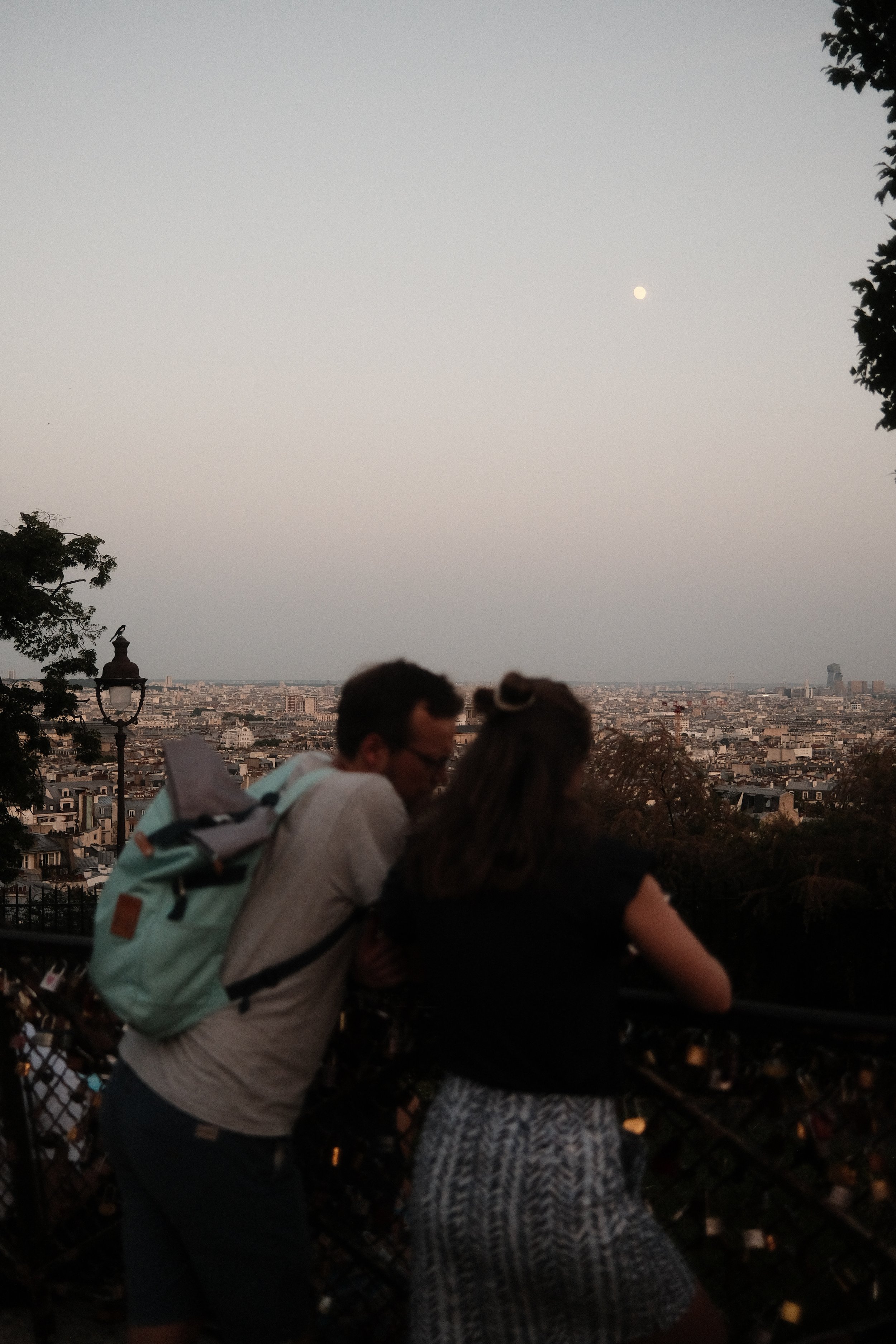 View of Paris skyline with the moon in the evening sky, people standing near a railing, some holding phones, trees and city buildings in the background.