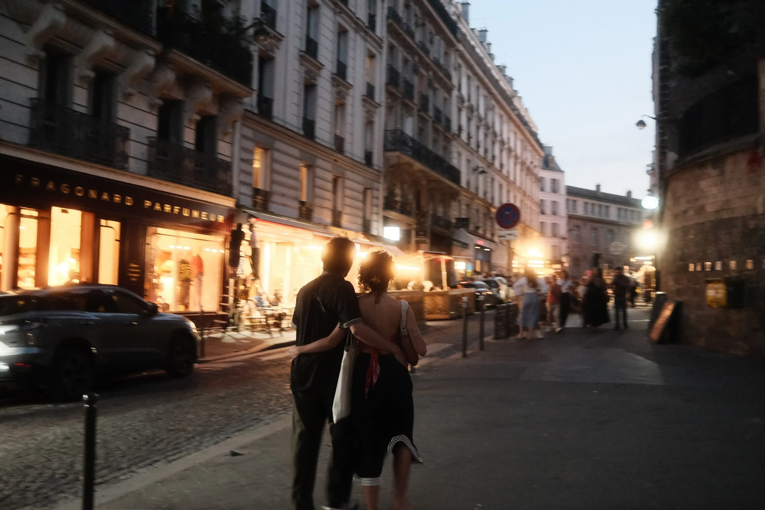 A couple walking arm in arm at dusk on a busy city street with buildings, shops, cars, and other pedestrians, illuminated by streetlights and shop windows.