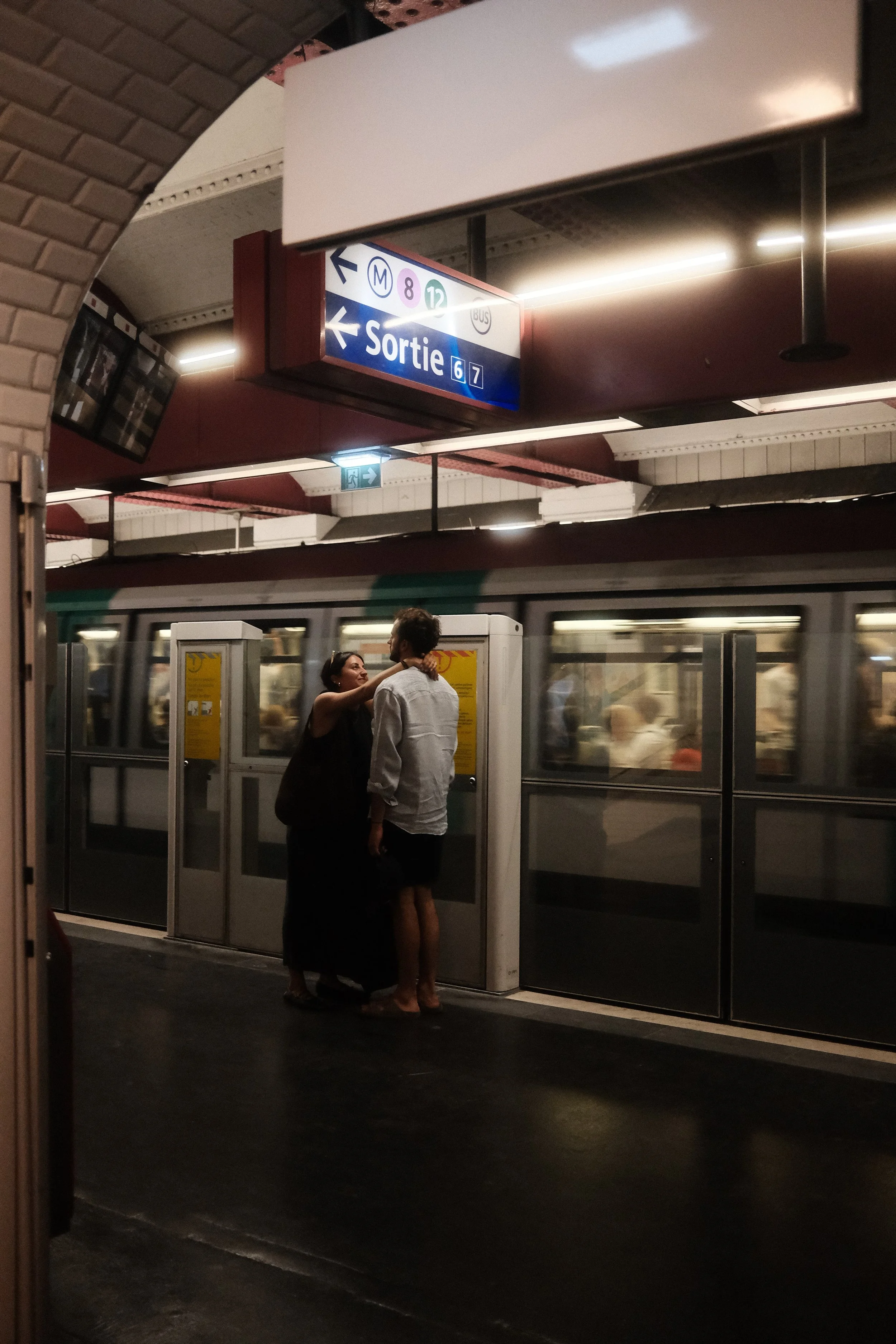 Two people are standing close together in a subway station, engaging in a conversation. A train is visible behind them, and there is a sign overhead indicating an exit and train routes.