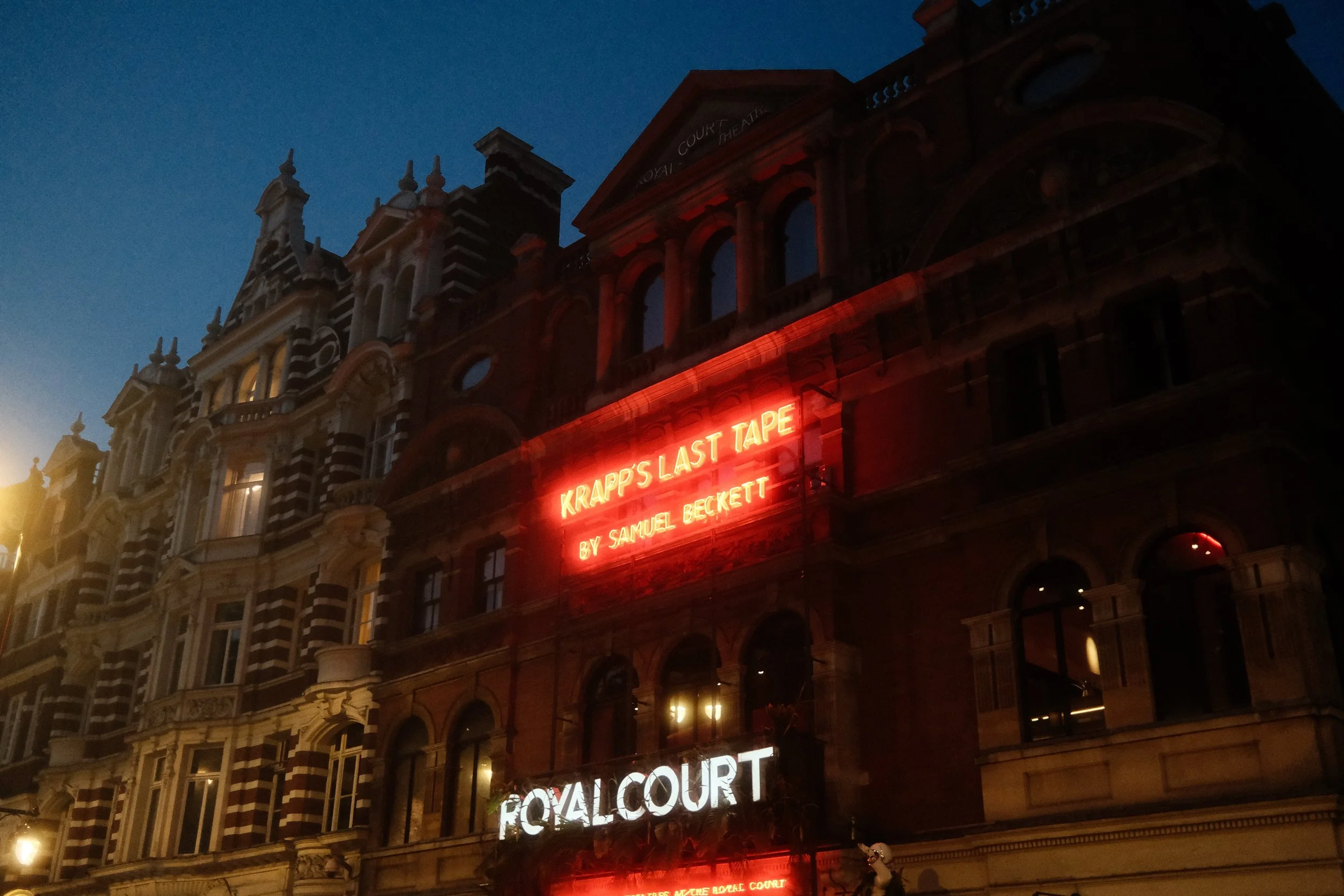 Nighttime view of a historic Victorian-style building with ornate architectural details, illuminated by a red neon sign for Krapp's Last Tape and other theater marquee lights.