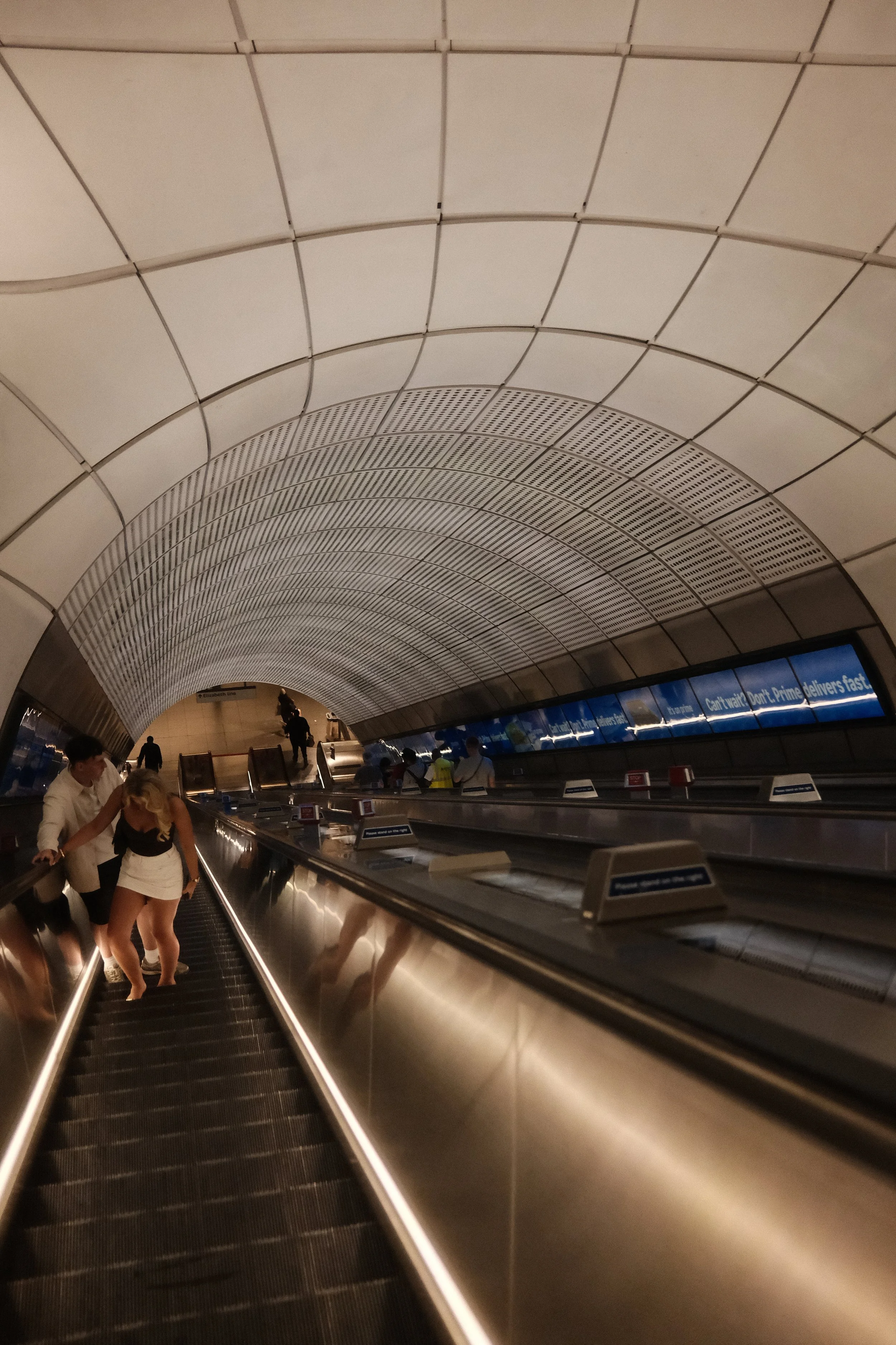 People using a moving walkway inside an airport terminal with a curved, paneled ceiling and digital advertisement screens.