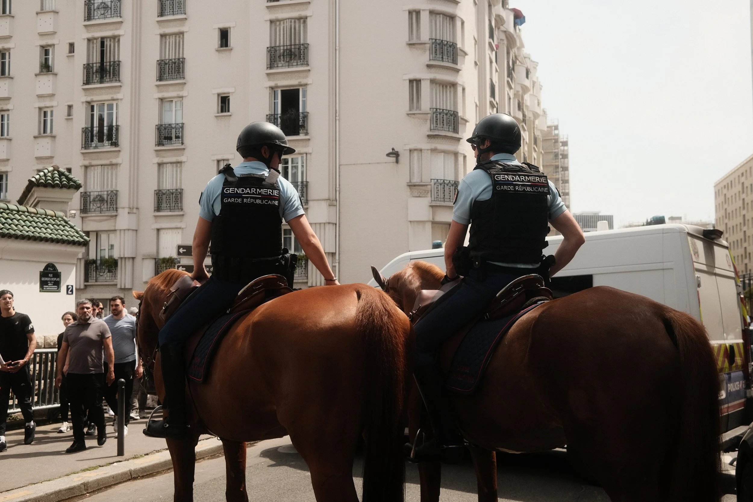 Two police officers on horseback patrol a city street, with a crowd of people and apartment buildings in the background.