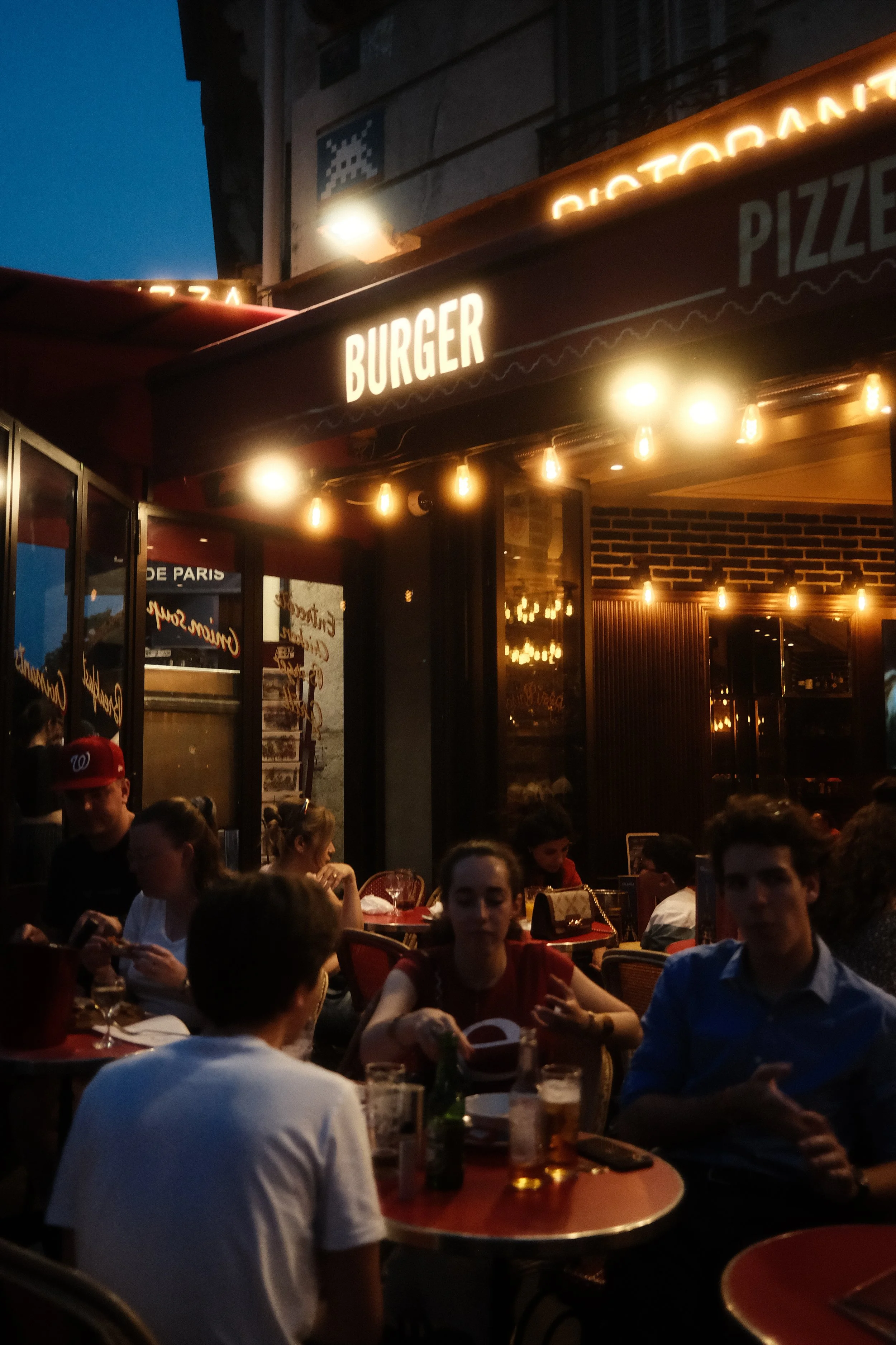 People dining inside a restaurant with a sign that reads 'BURGER' above the entrance, warm lighting, and a mix of men and women sitting at tables.