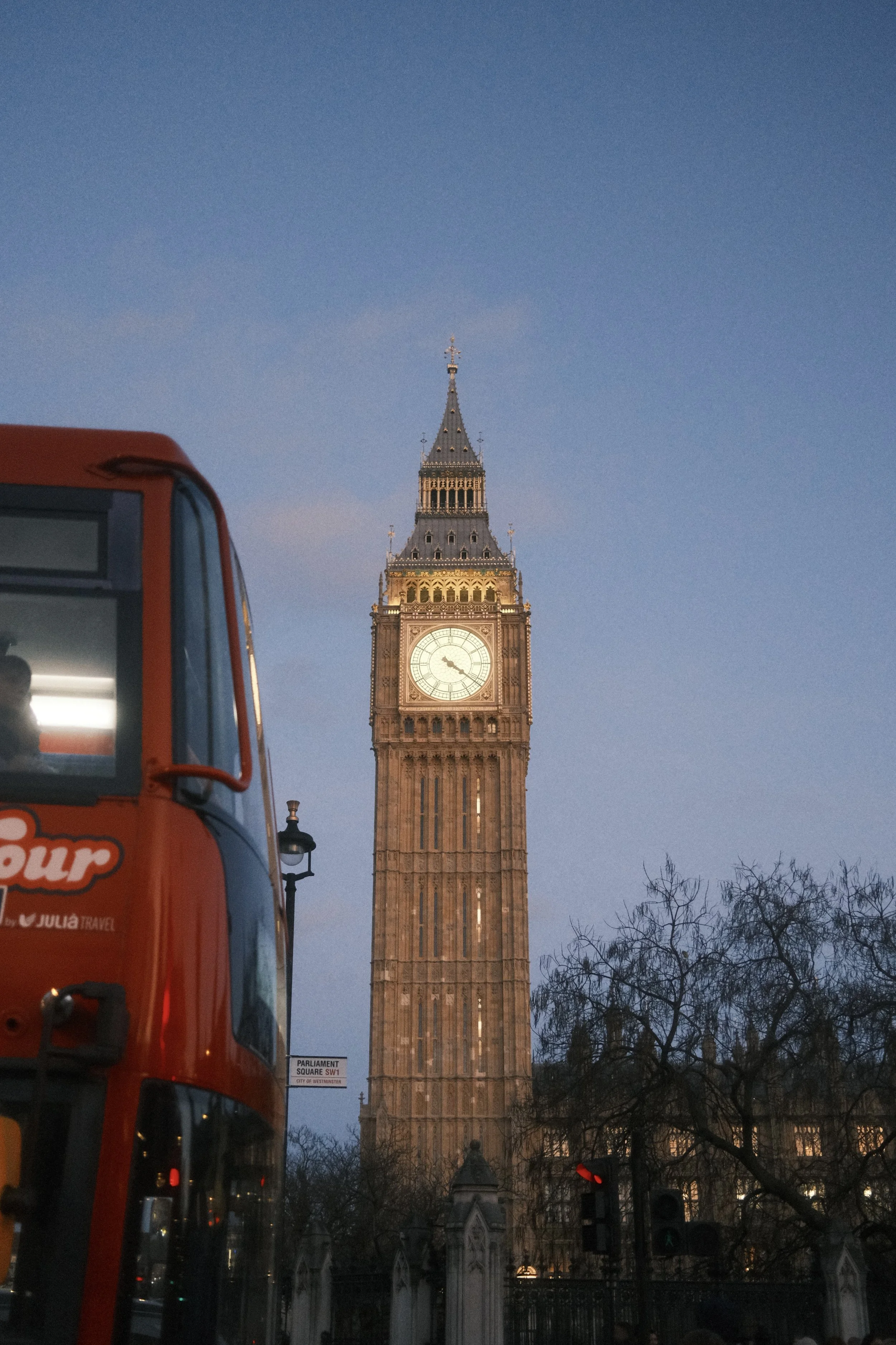 A view of Big Ben clock tower in London, England, during dusk with a red double-decker bus in the foreground and leafless trees along the street.