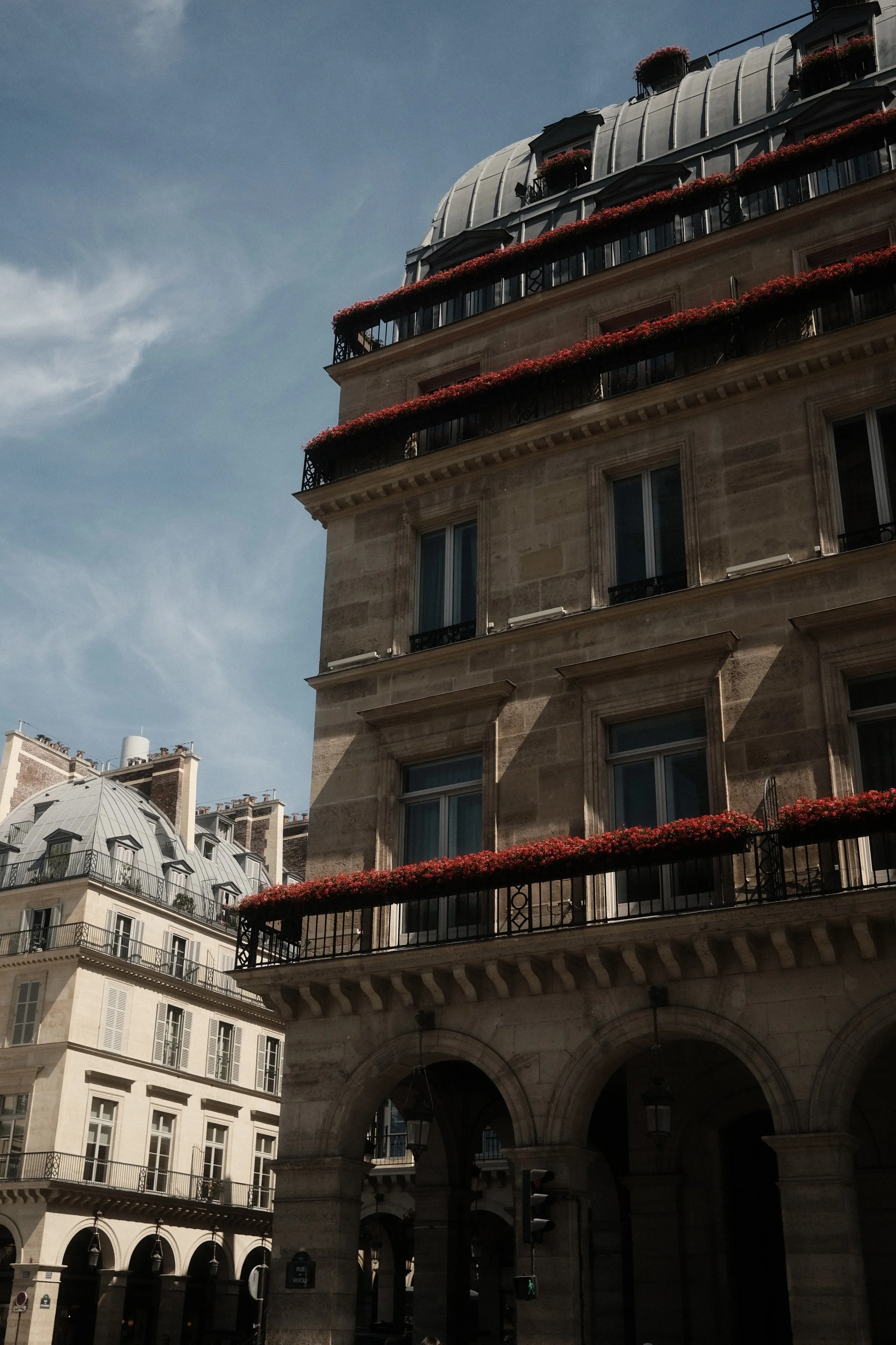 City street scene featuring historic buildings with wrought-iron balconies and red flower boxes, stone archway, and a traffic light with pedestrian signal, under a partly cloudy sky.