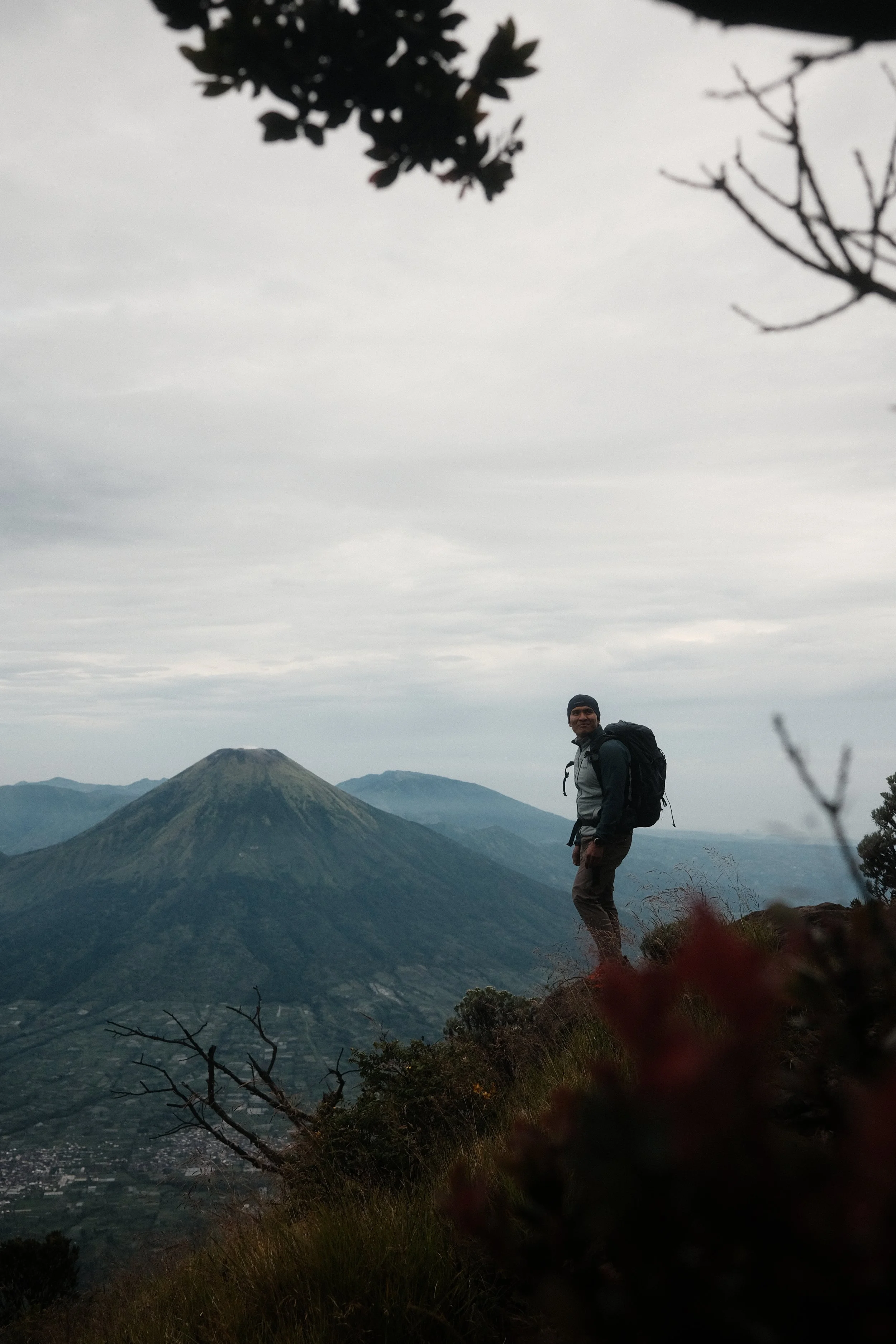 A hiker with a backpack standing on a mountain trail during overcast weather, with a volcanic mountain in the background and trees in the foreground.