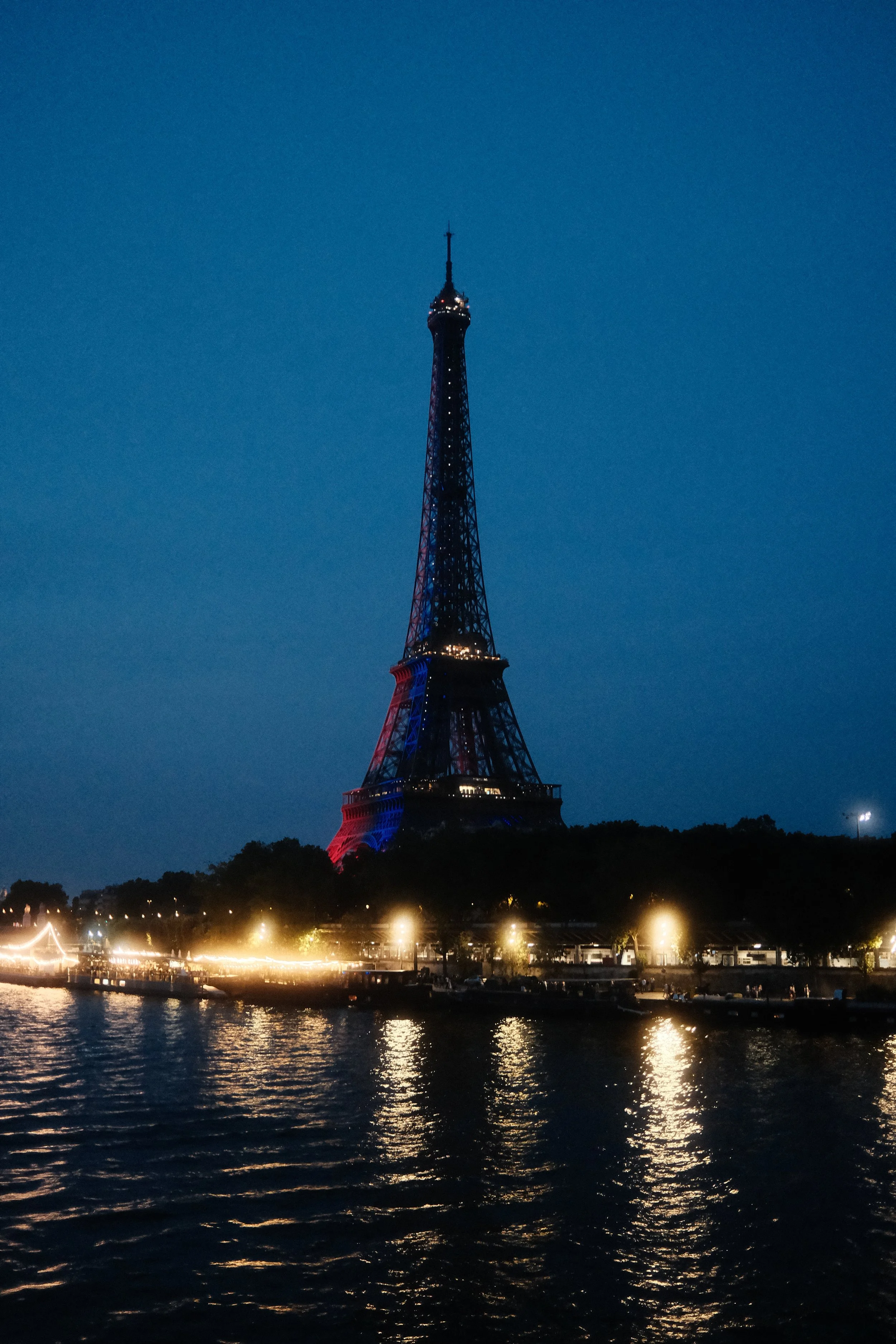 Night view of the Eiffel Tower illuminated with red, white, and blue lights, situated along a river with reflections of the lights on the water and a clear dark sky overhead.