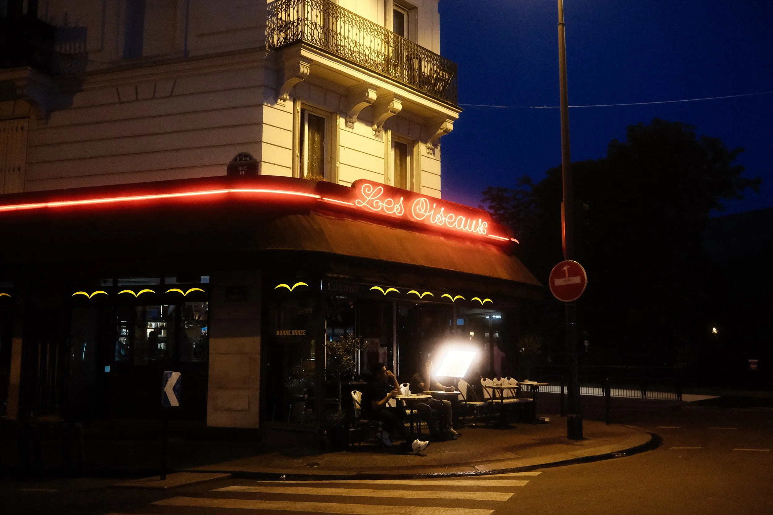 Nighttime street scene with a corner building featuring a balcony and a neon sign that reads 'Les Oiseaux'. The storefront below has a red awning, and several tables and chairs are outside with people sitting beneath streetlights.