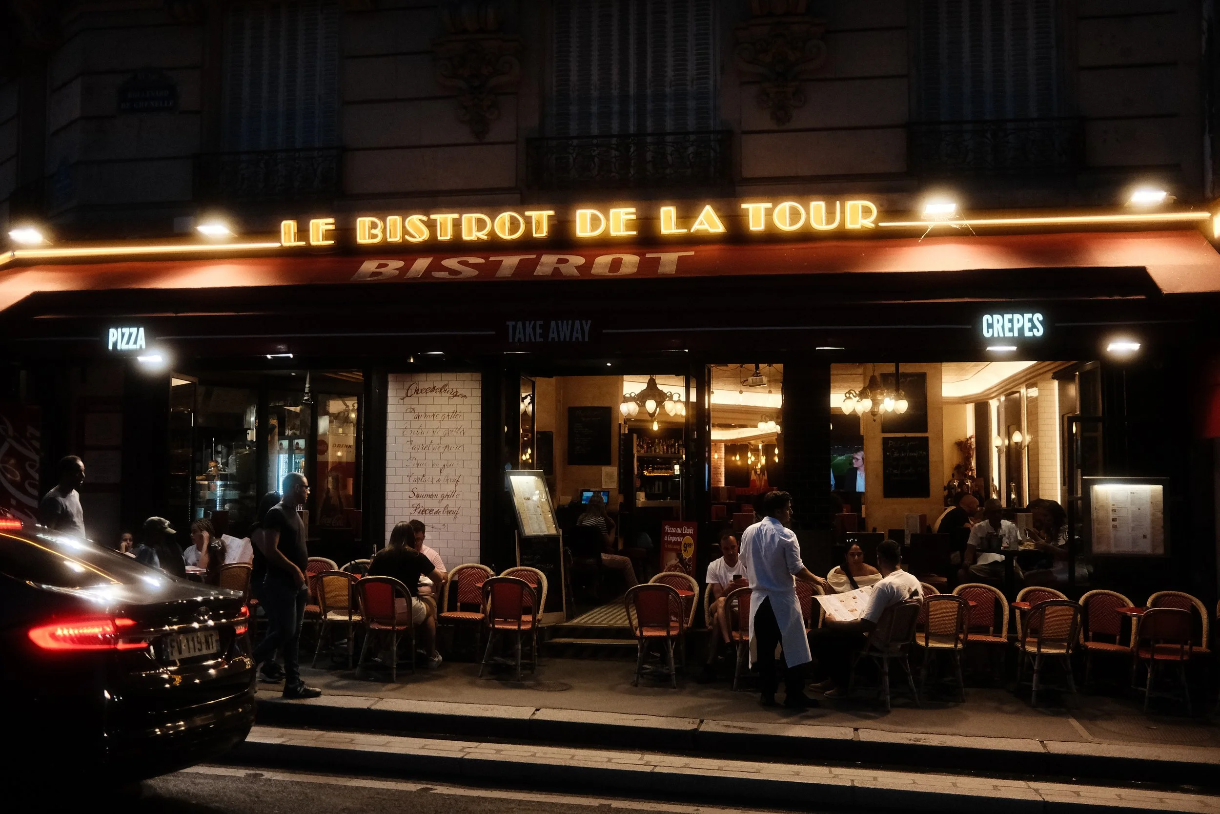 Night view of a restaurant named 'Le Bistrot de La Tour' with neon signs, outdoor seating, and people dining, streetlights illuminating the sidewalk, and a parked car in the foreground.