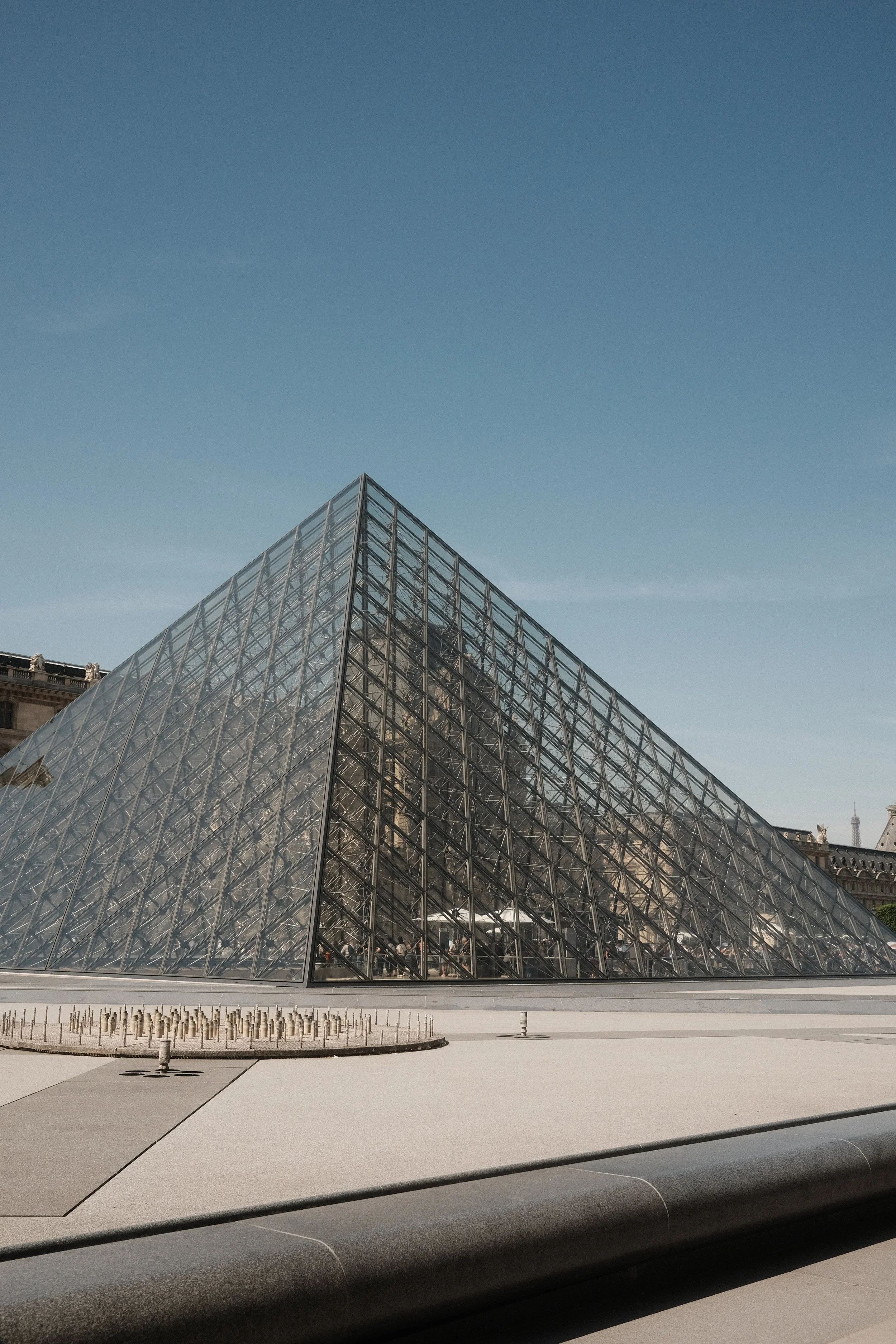 The Louvre Pyramid, a glass and metal structure located in the courtyard of the Louvre Museum in Paris, France, with a clear blue sky above.