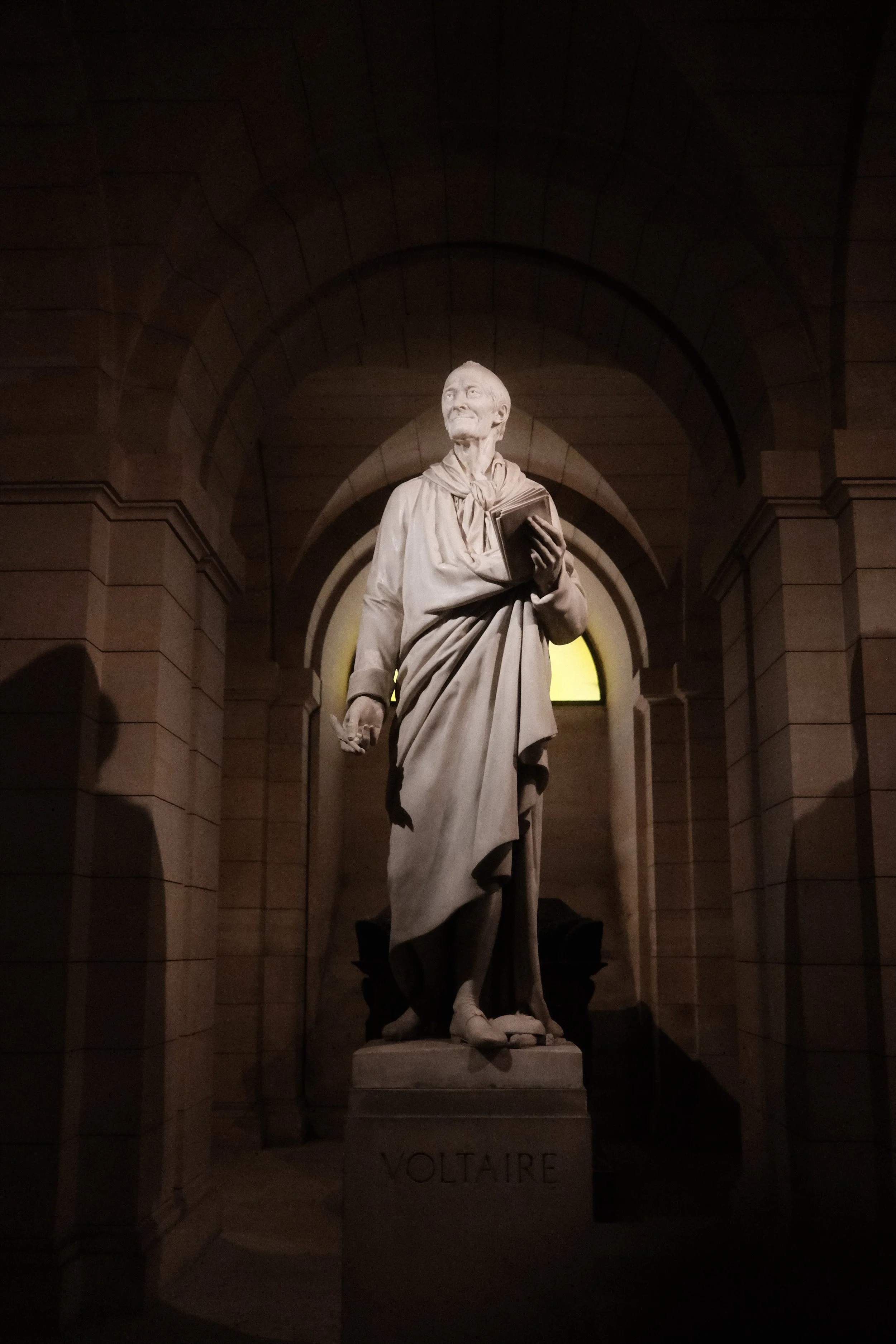 Statue of Voltaire in a dimly lit indoor setting with stone arches in the background.