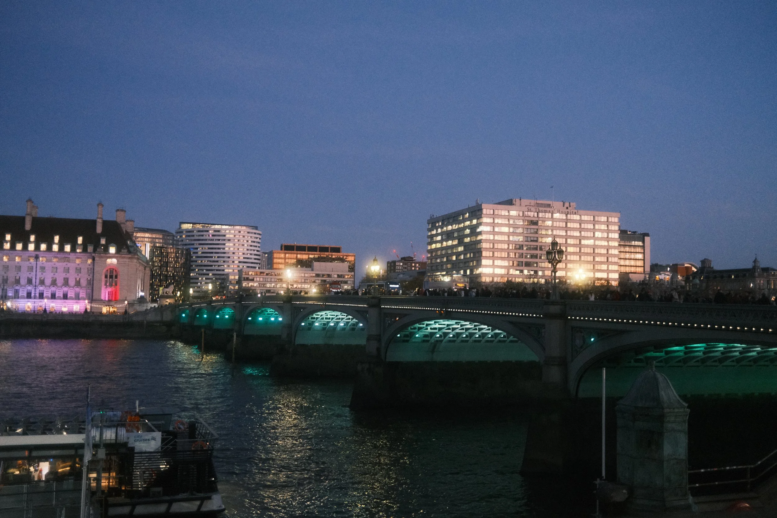 Nighttime cityscape with illuminated buildings, a bridge with green-lit arches, and a river reflecting city lights.