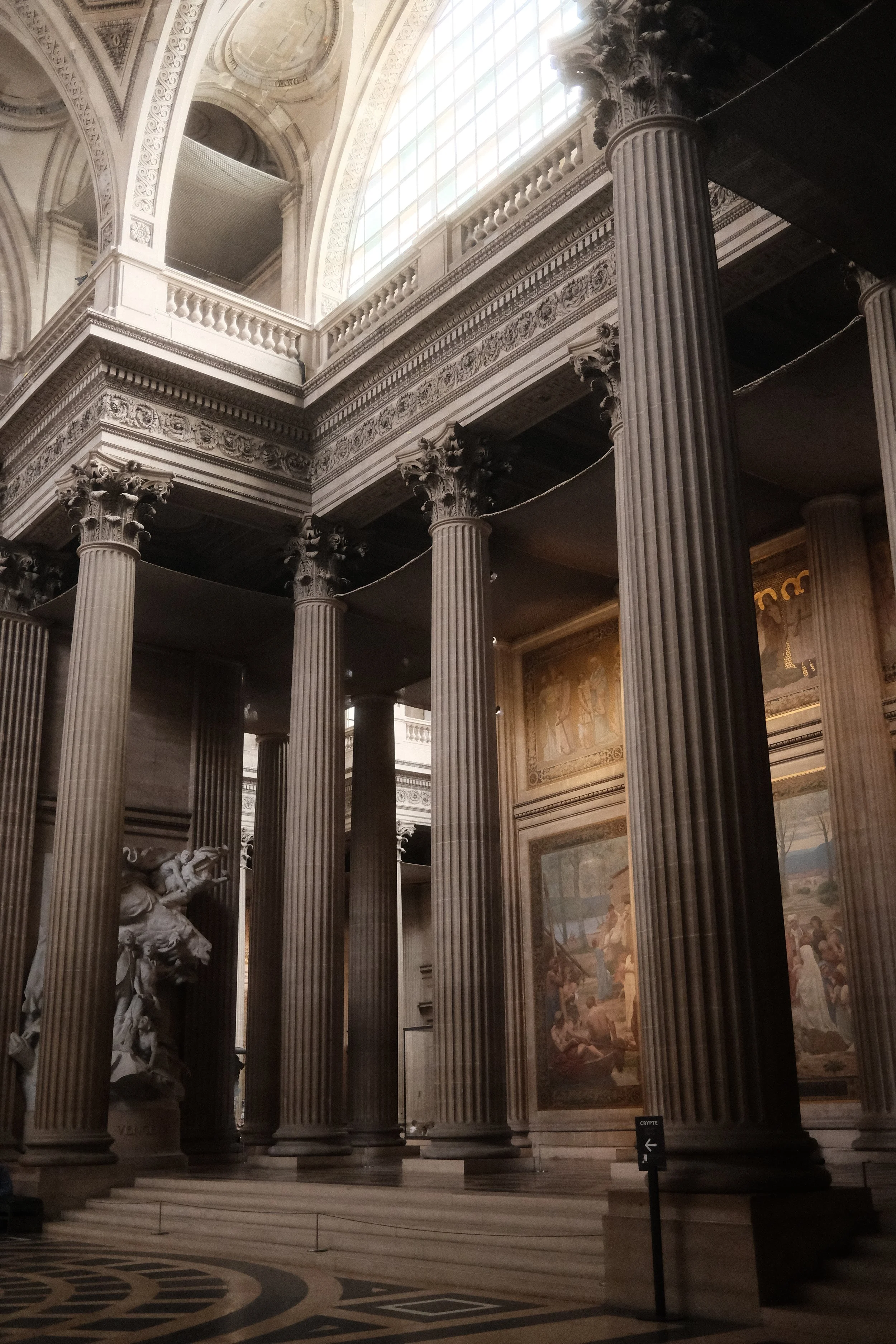 Interior of a grand building with tall, fluted columns with ornate Corinthian capitals, detailed friezes, large windows, and classical murals or paintings on the walls.