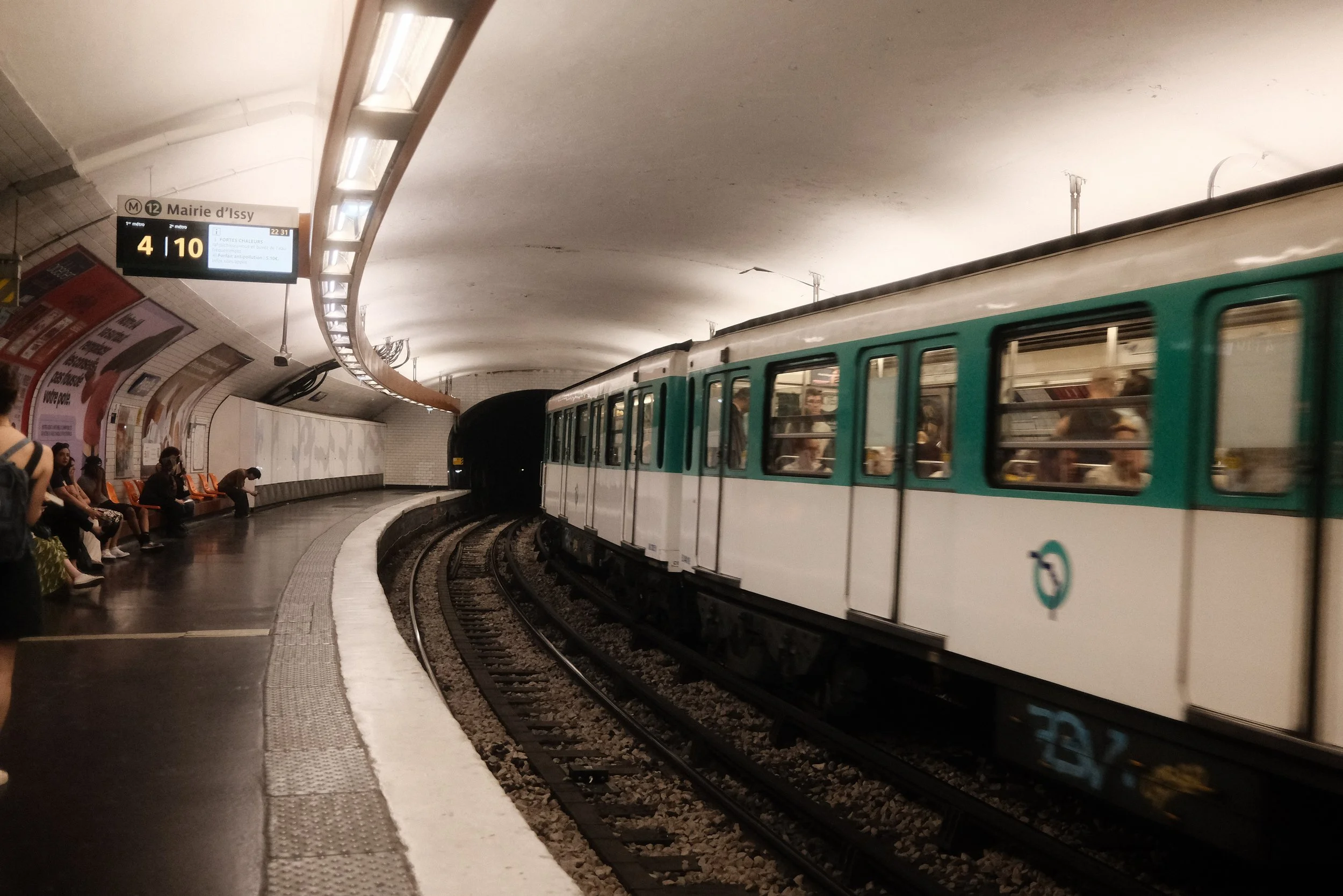 An underground metro station with a train arriving or departing, passengers sitting on benches waiting, and a digital sign displaying metro line information.