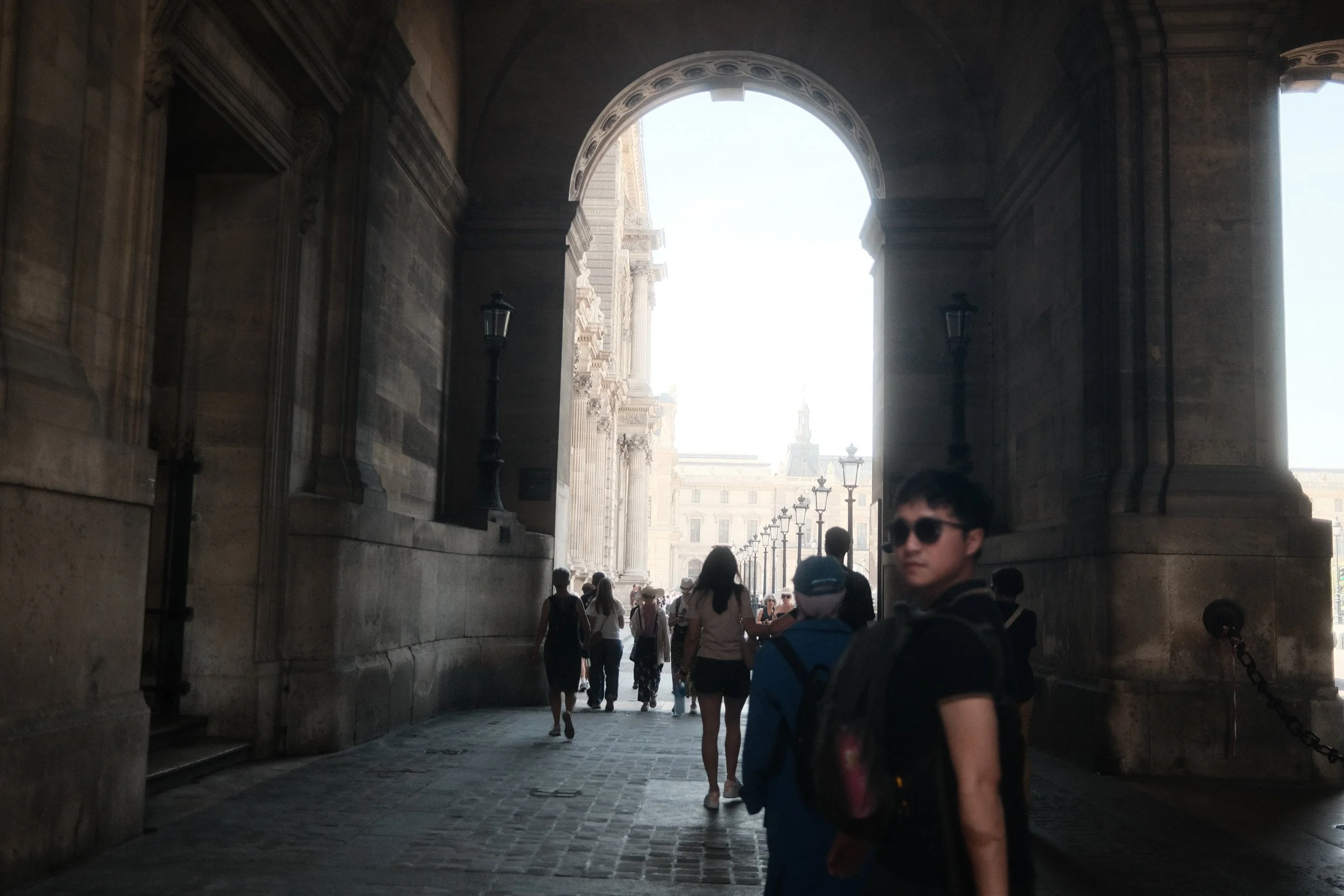 People walking through an archway with a sunny cityscape in the background.
