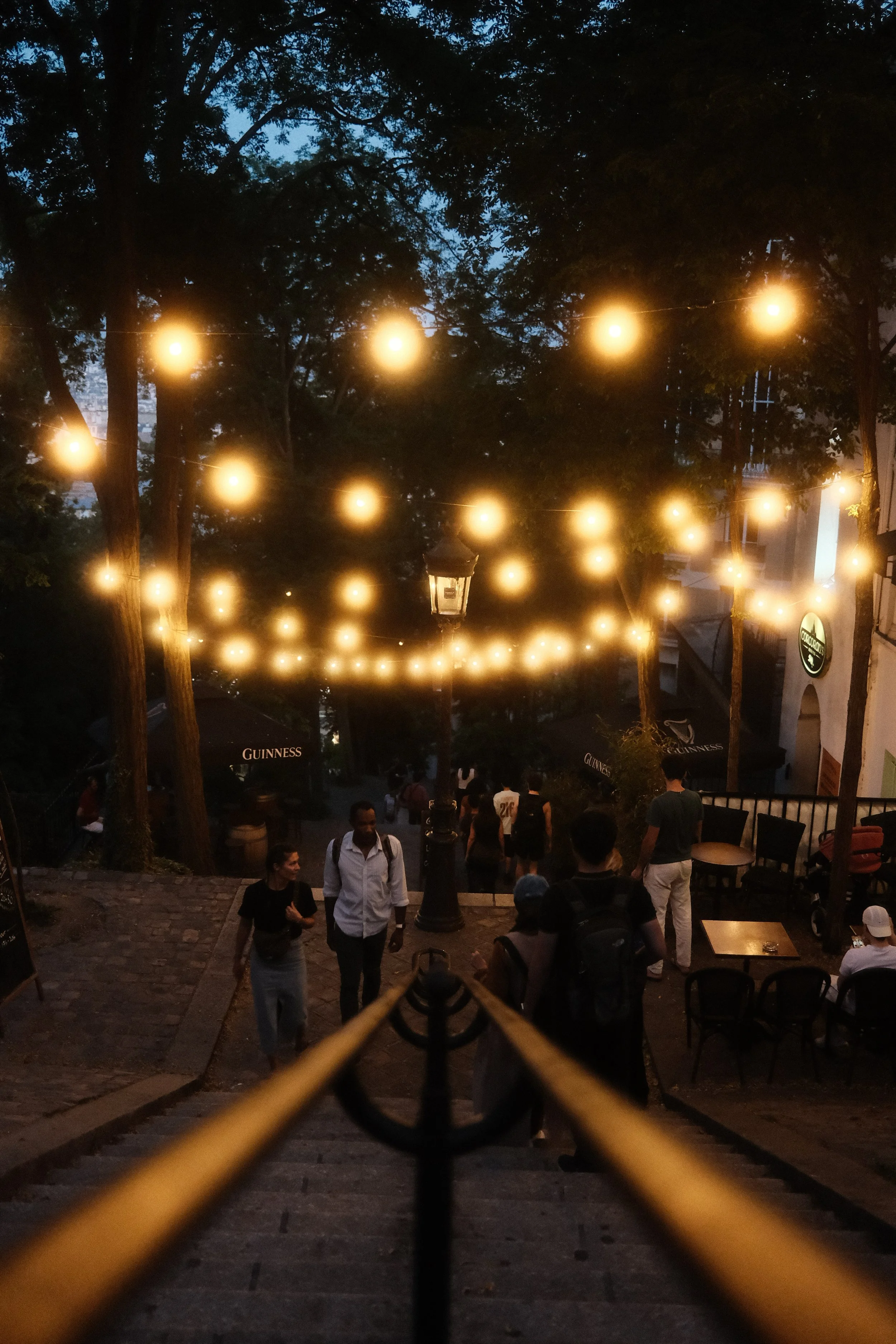 An outdoor gathering with string lights hanging between trees during dusk, with people walking and sitting in a patio area.