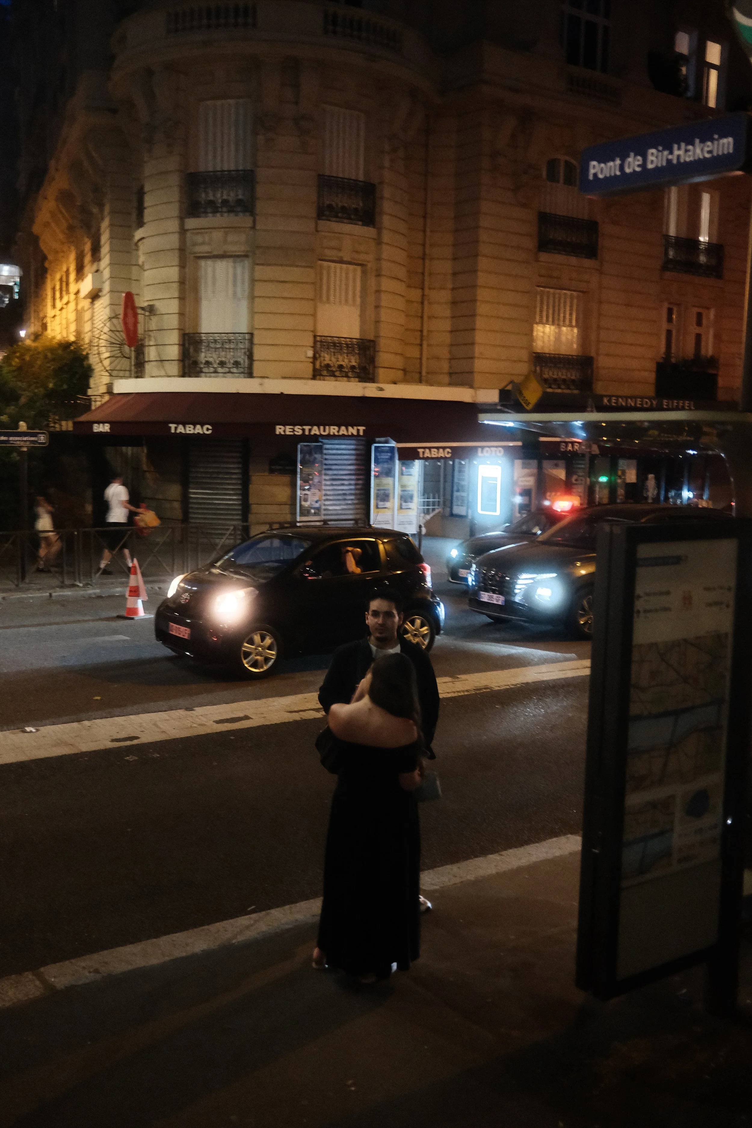 Nighttime street scene with a building featuring classic architecture, several parked cars on the street, and people standing at the sidewalk. A street sign reading 'Pont de Bir-Hakeim' is visible along with a small illuminated kiosk and a building w