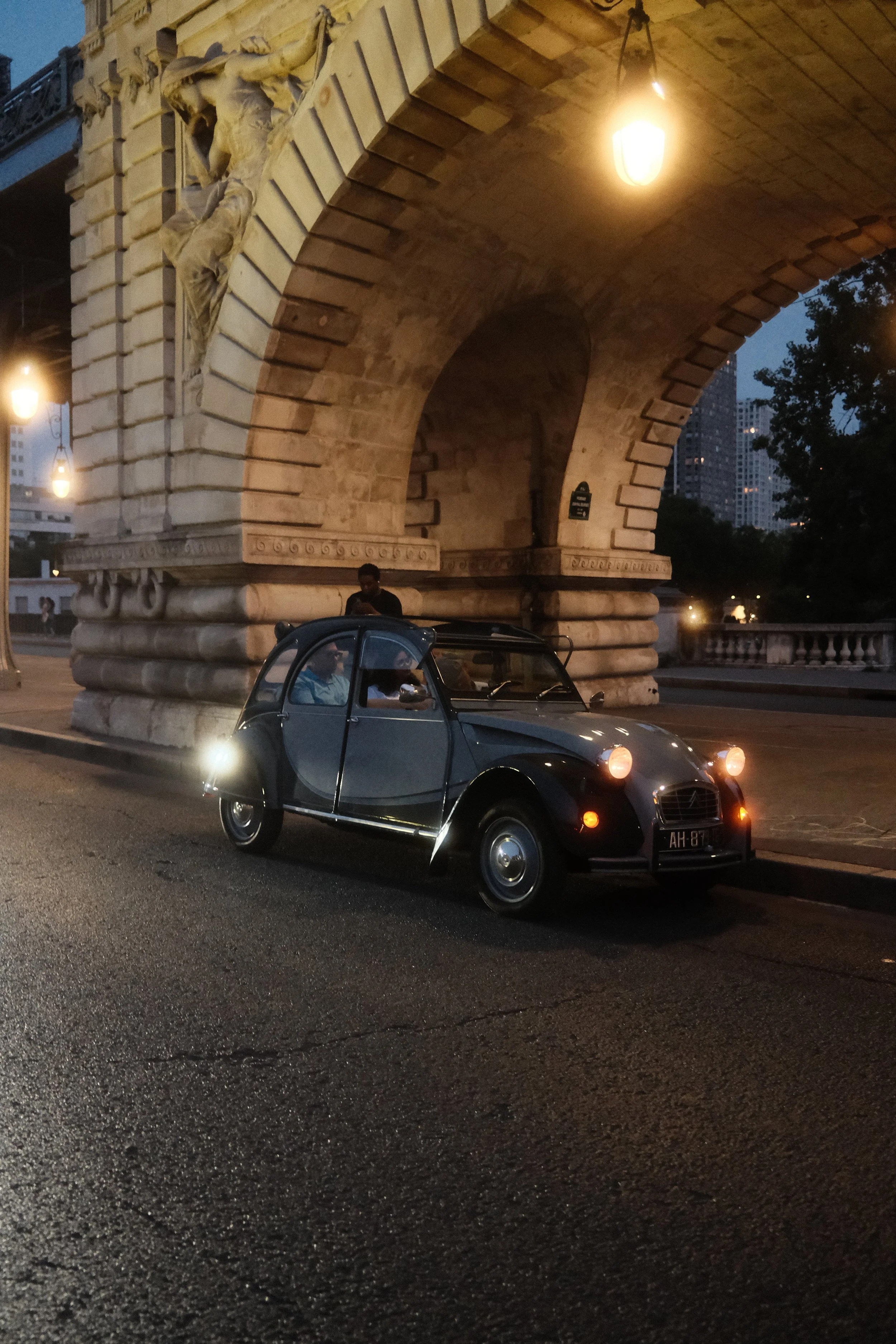 A vintage black car with its headlights on driving under a large, illuminated stone arch bridge. The scene is at dusk or early evening, with city buildings and trees visible in the background.