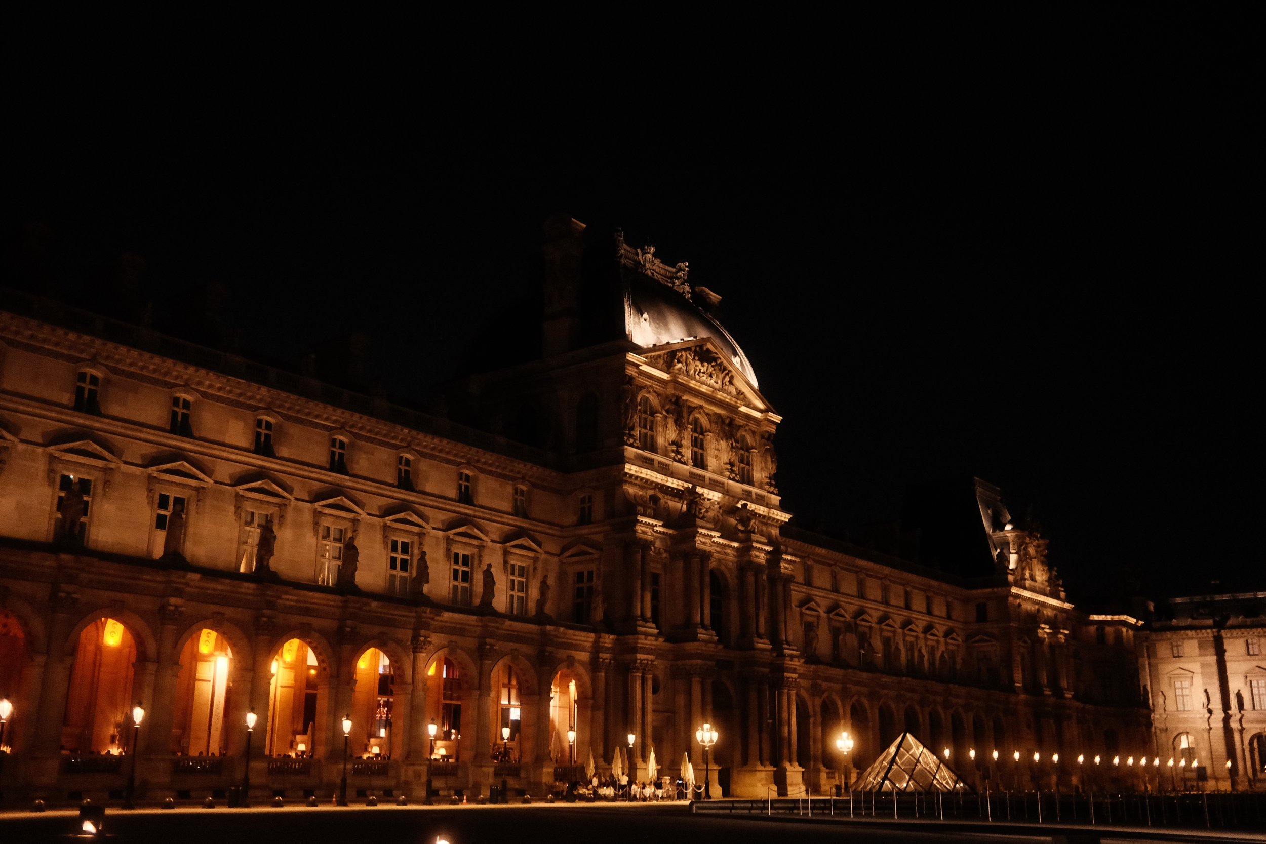 Night view of a historic building with ornate architecture, illuminated from inside with warm lights, and a glass pyramid structure in front.