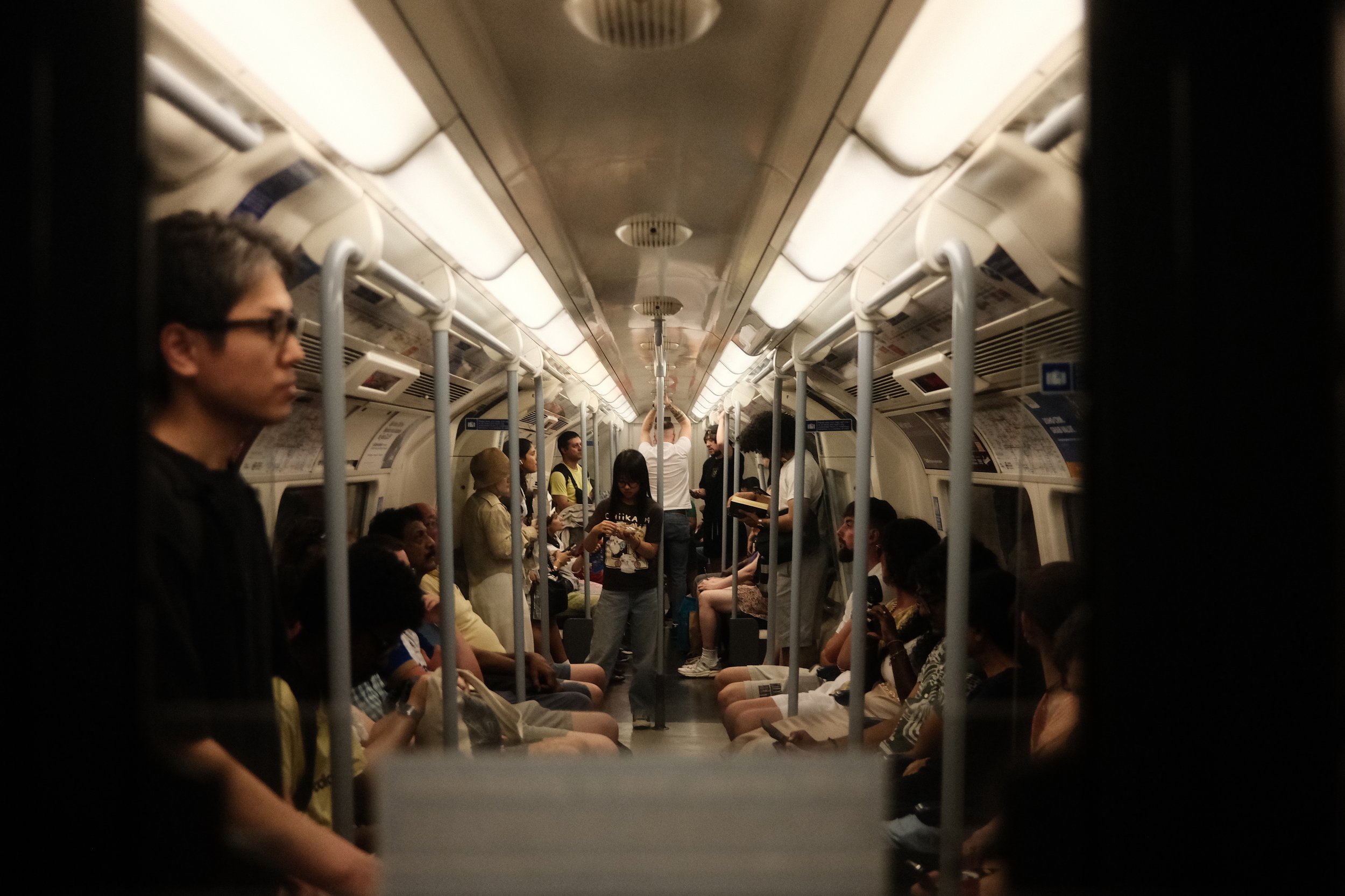 View of the interior of a subway train with passengers seated and standing, seen through a window or opening with vertical bars. People are engaged in various activities, including using phones and conversing.