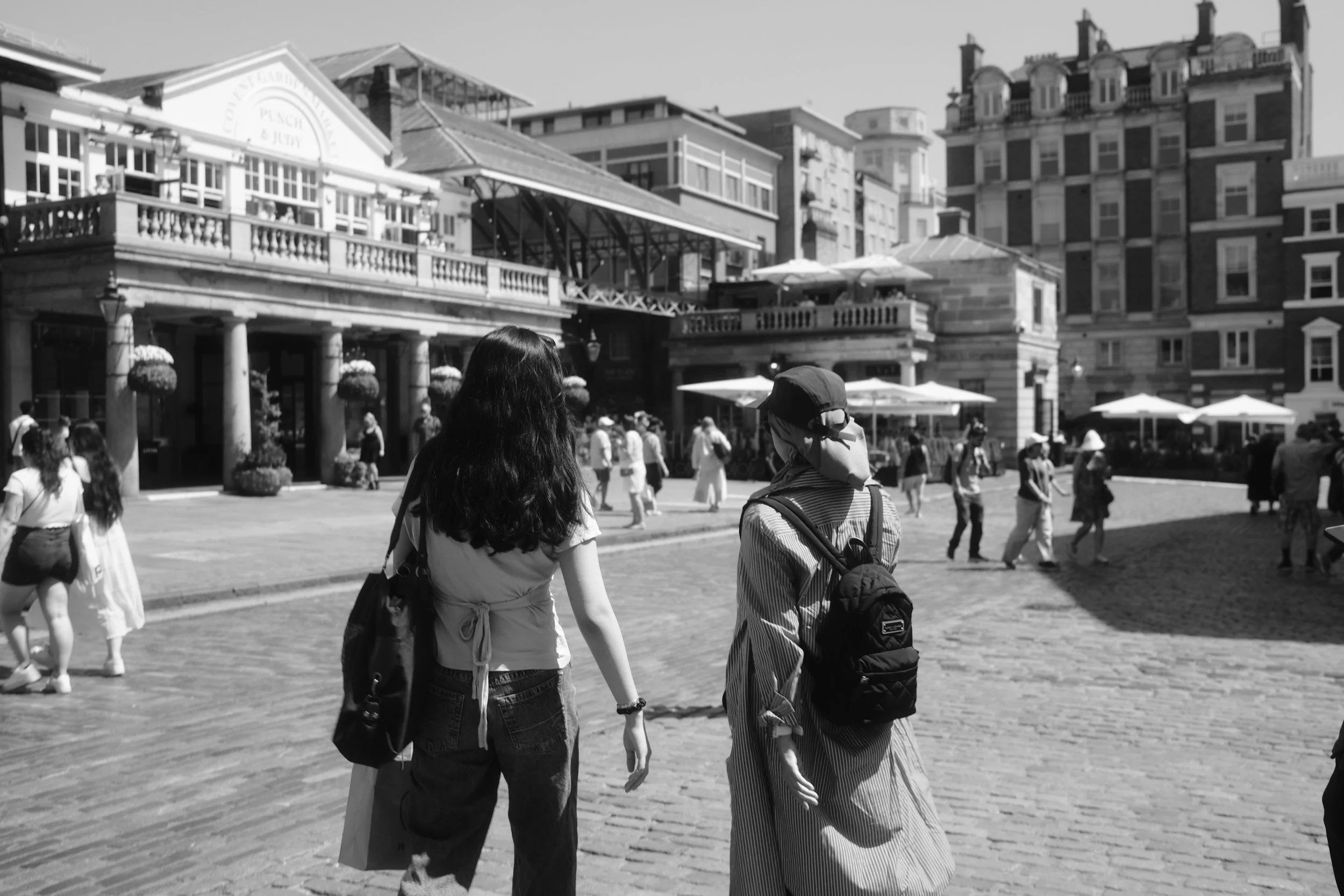 People walking in an outdoor plaza area with historic buildings and outdoor seating with umbrellas in the background