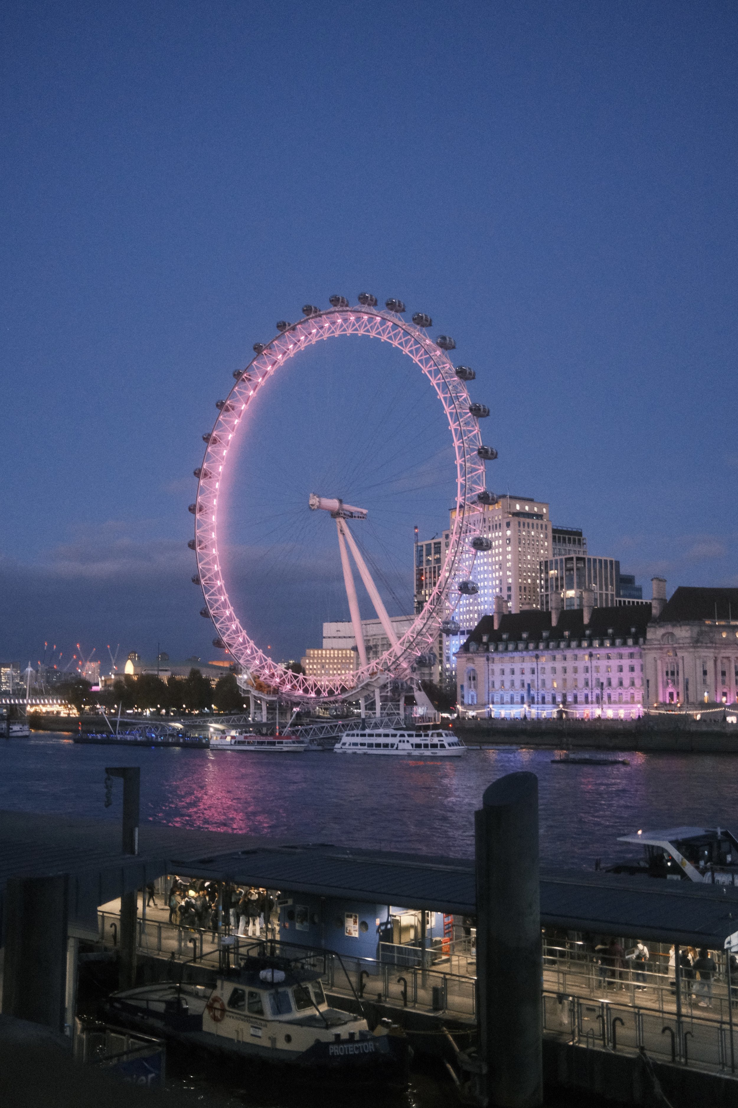 Nighttime view of the London Eye Ferris wheel illuminated in pink, located on the South Bank of the River Thames with surrounding buildings and boats reflecting on the water.