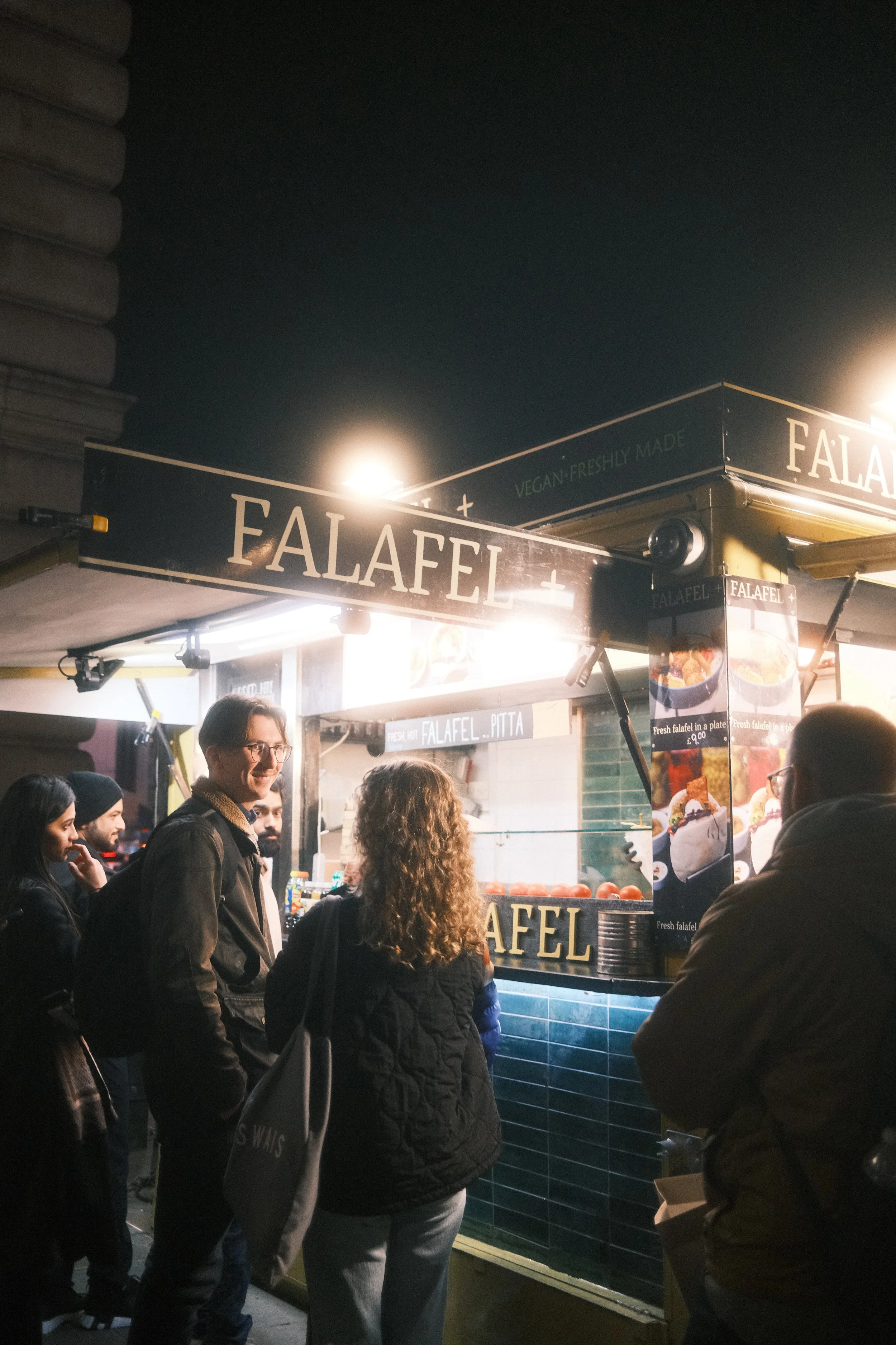 Night scene of a food stall selling falafel, with people standing in line and the seller inside, illuminated by bright lights.