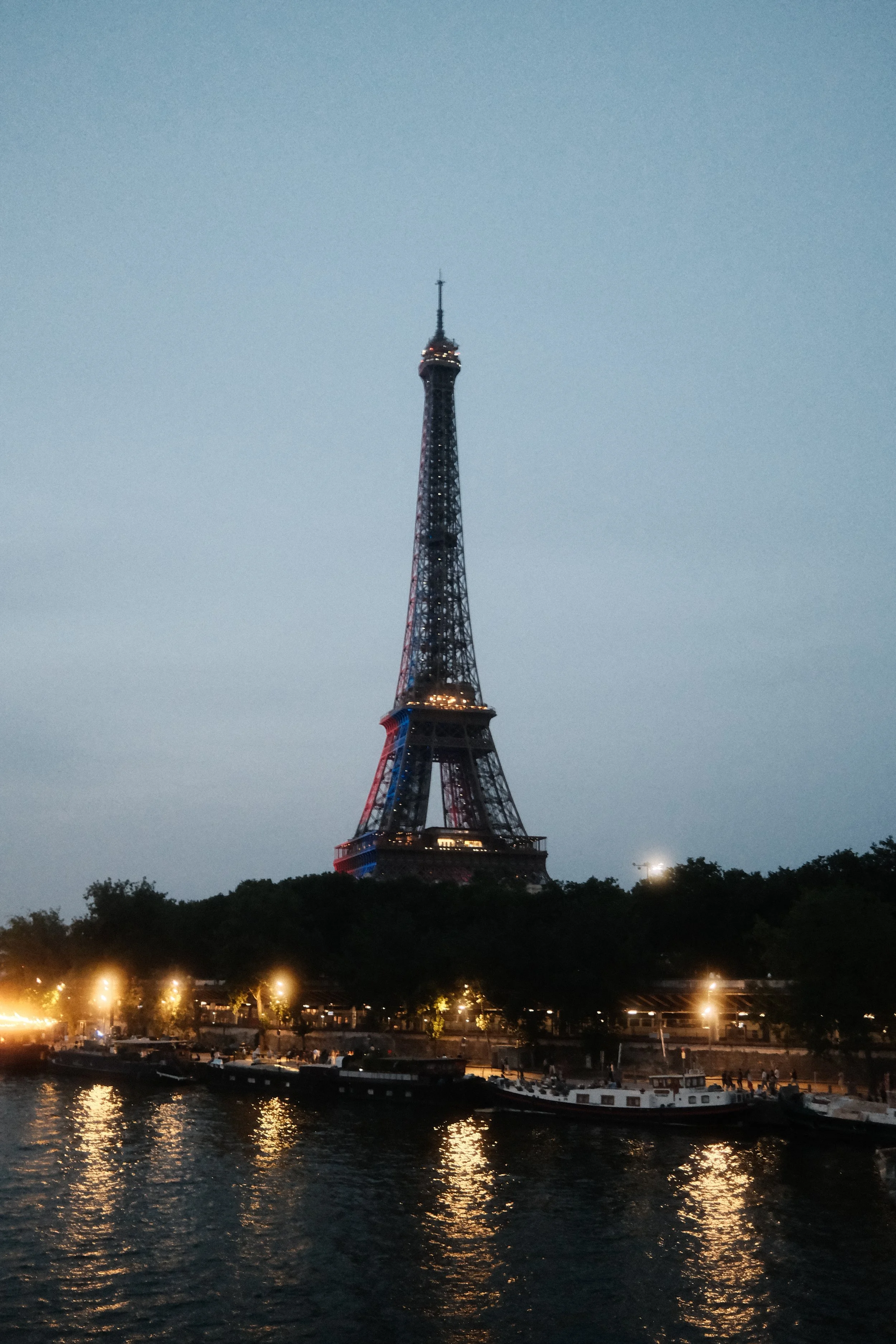 The Eiffel Tower illuminated with colorful lights at dusk, viewed from across a river with boats and a small dock in the foreground.
