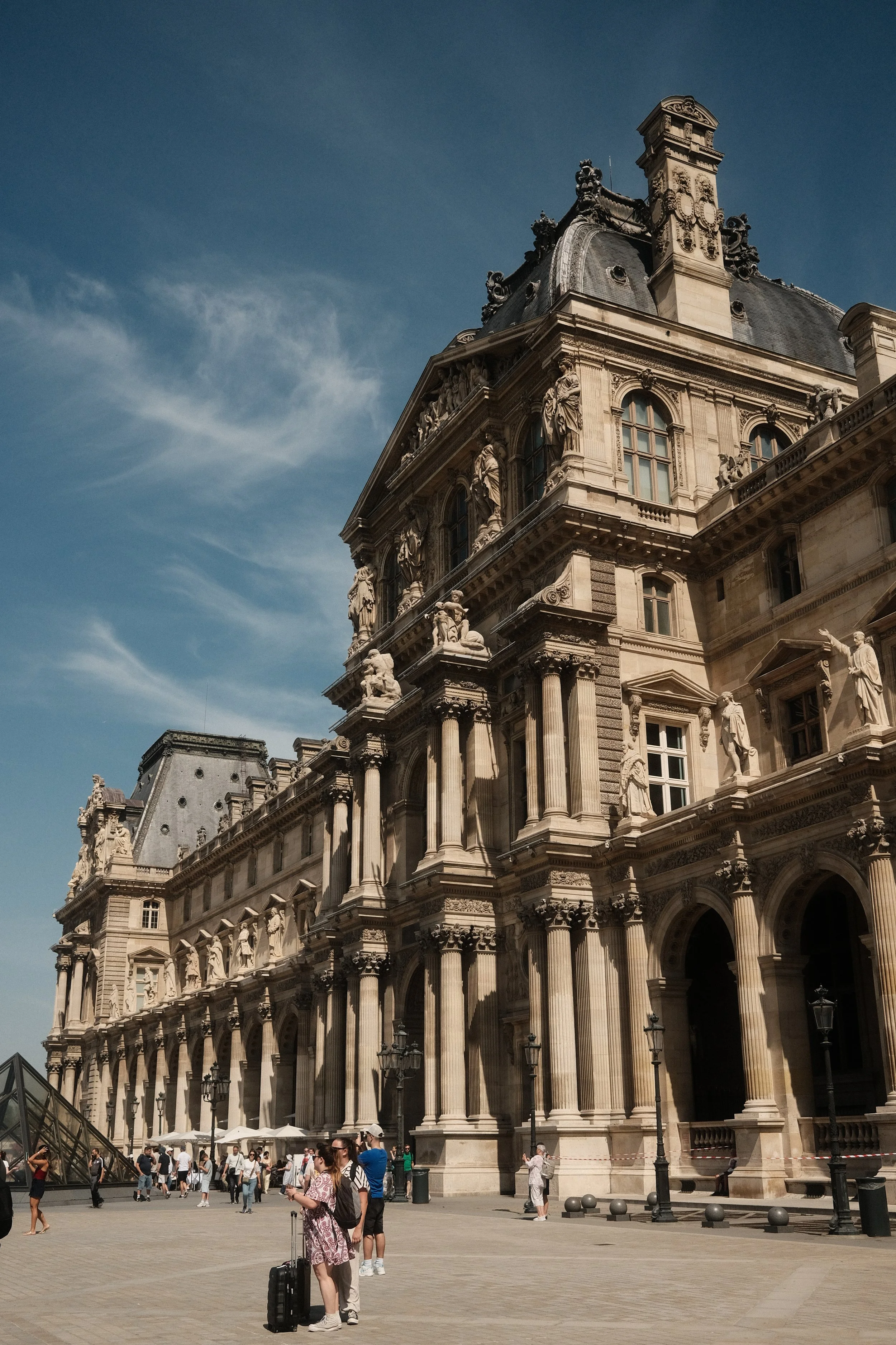 People walking and standing outside a historic, ornate building with statues and columns, under a clear blue sky.