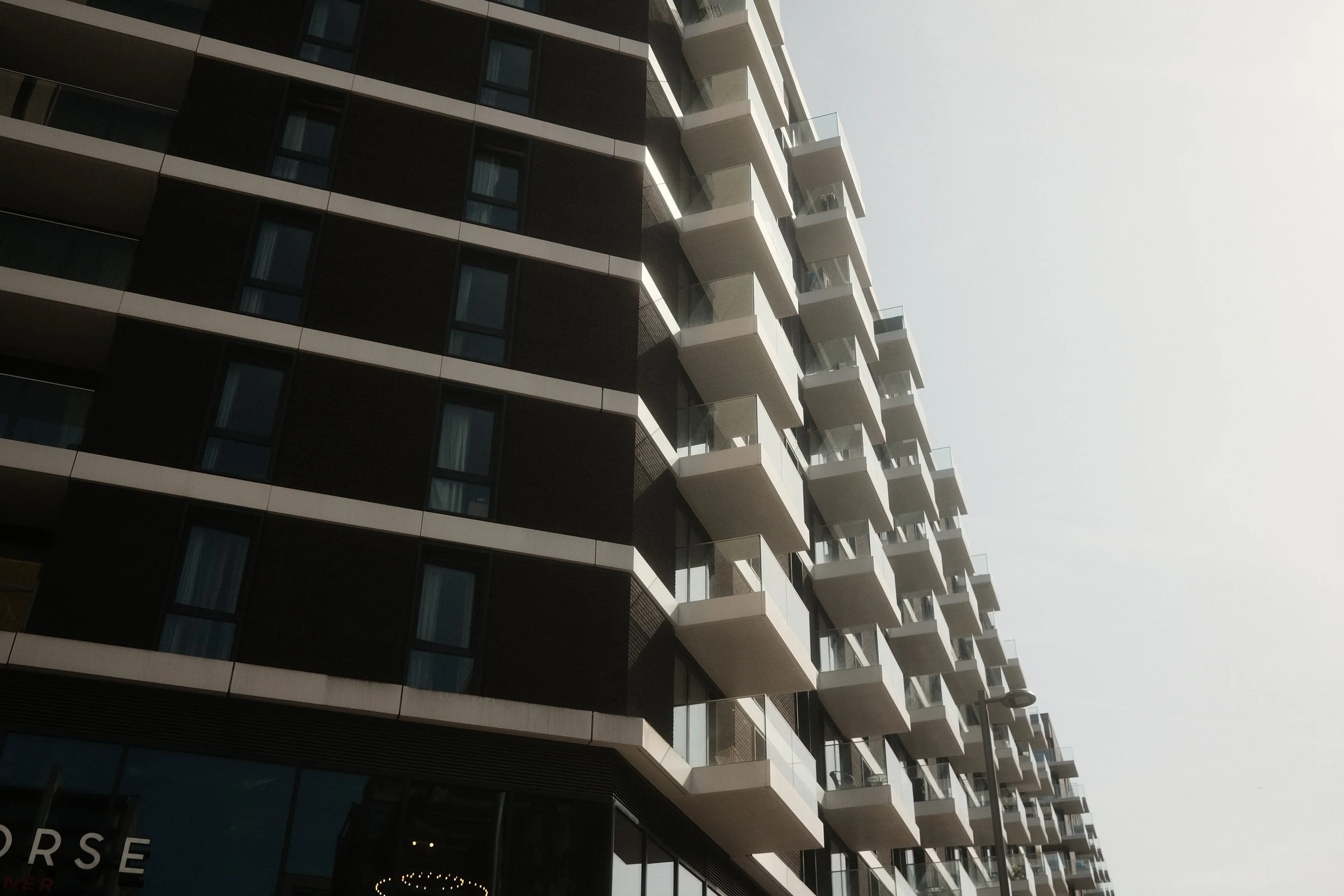 A modern multi-story building with balconies and glass railings, viewed from a low angle against a clear sky.