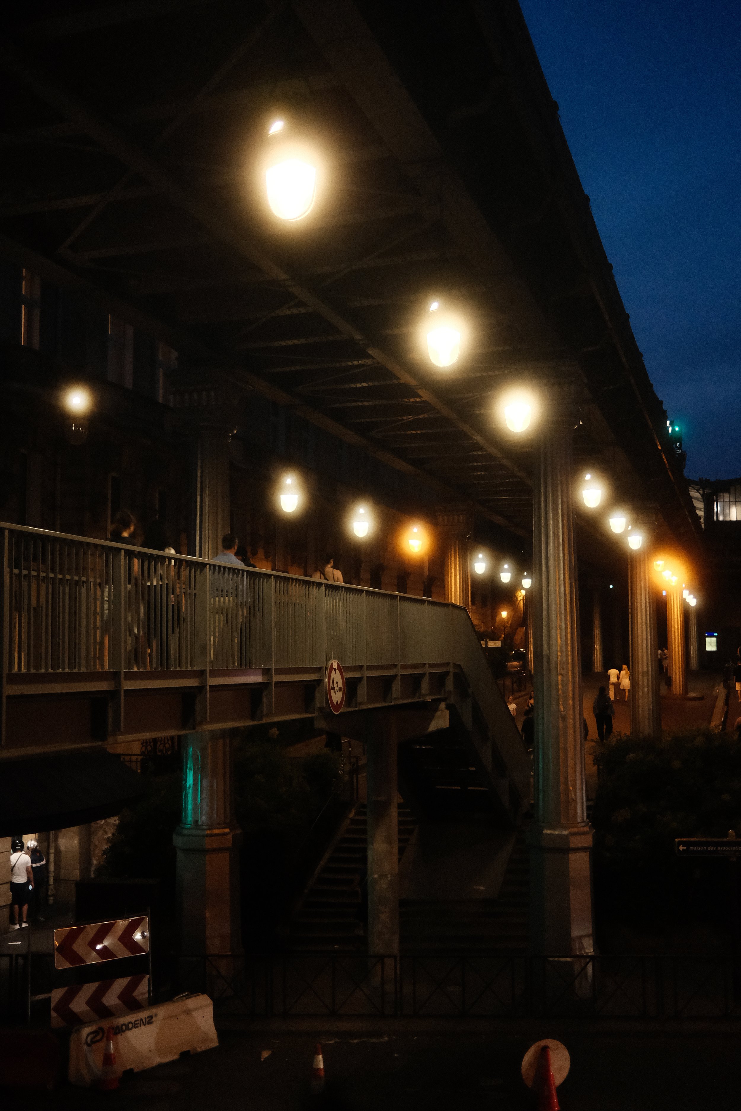 Nighttime scene of an elevated railway track with bright lights underneath. Several people are walking underneath, and there are construction cones and barriers in the foreground. The background features dark blue sky and some city lights.