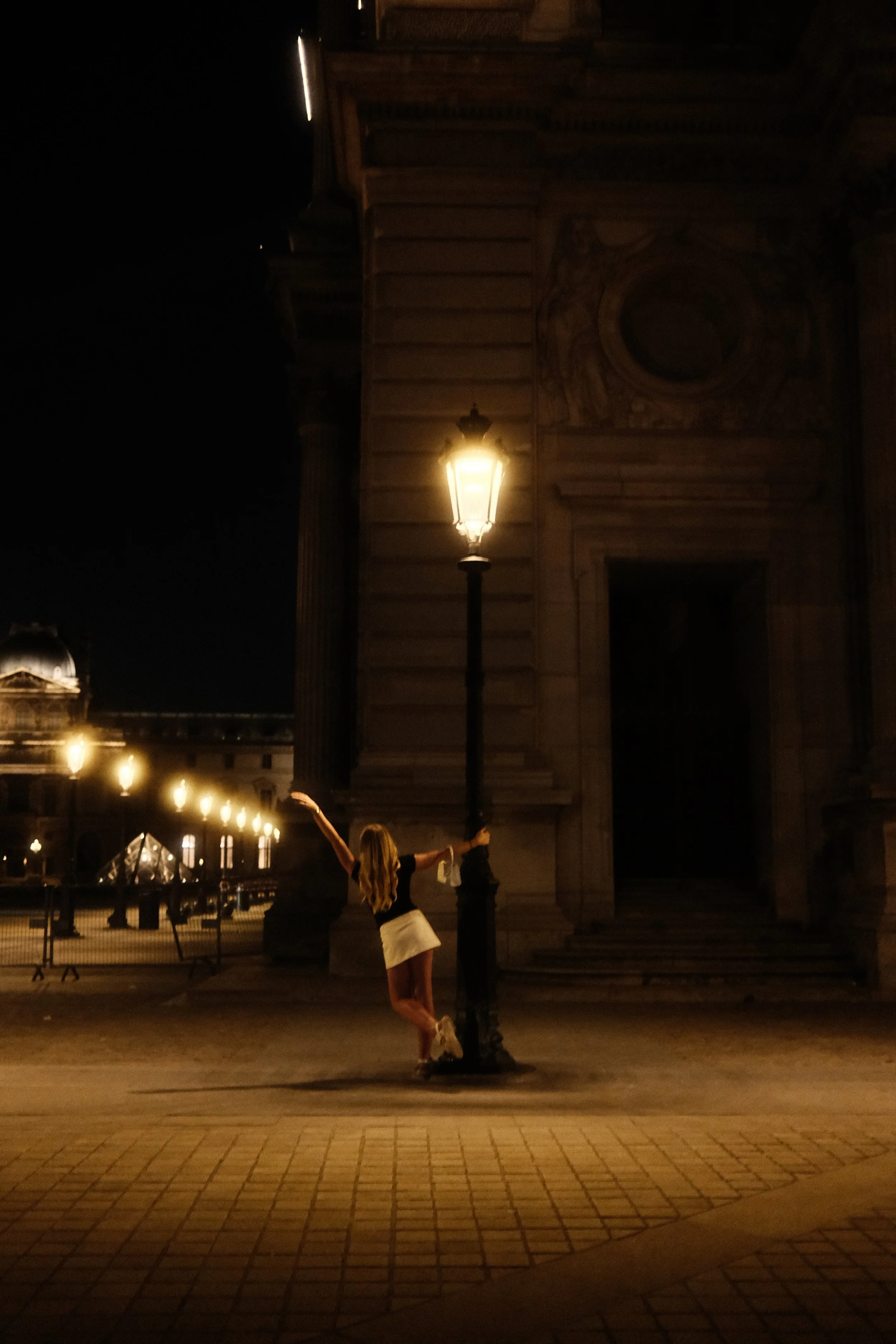 A woman in a black top and white skirt with long blonde hair posing playfully beside a streetlamp at night outside an ornate building with sculptures and architectural details.