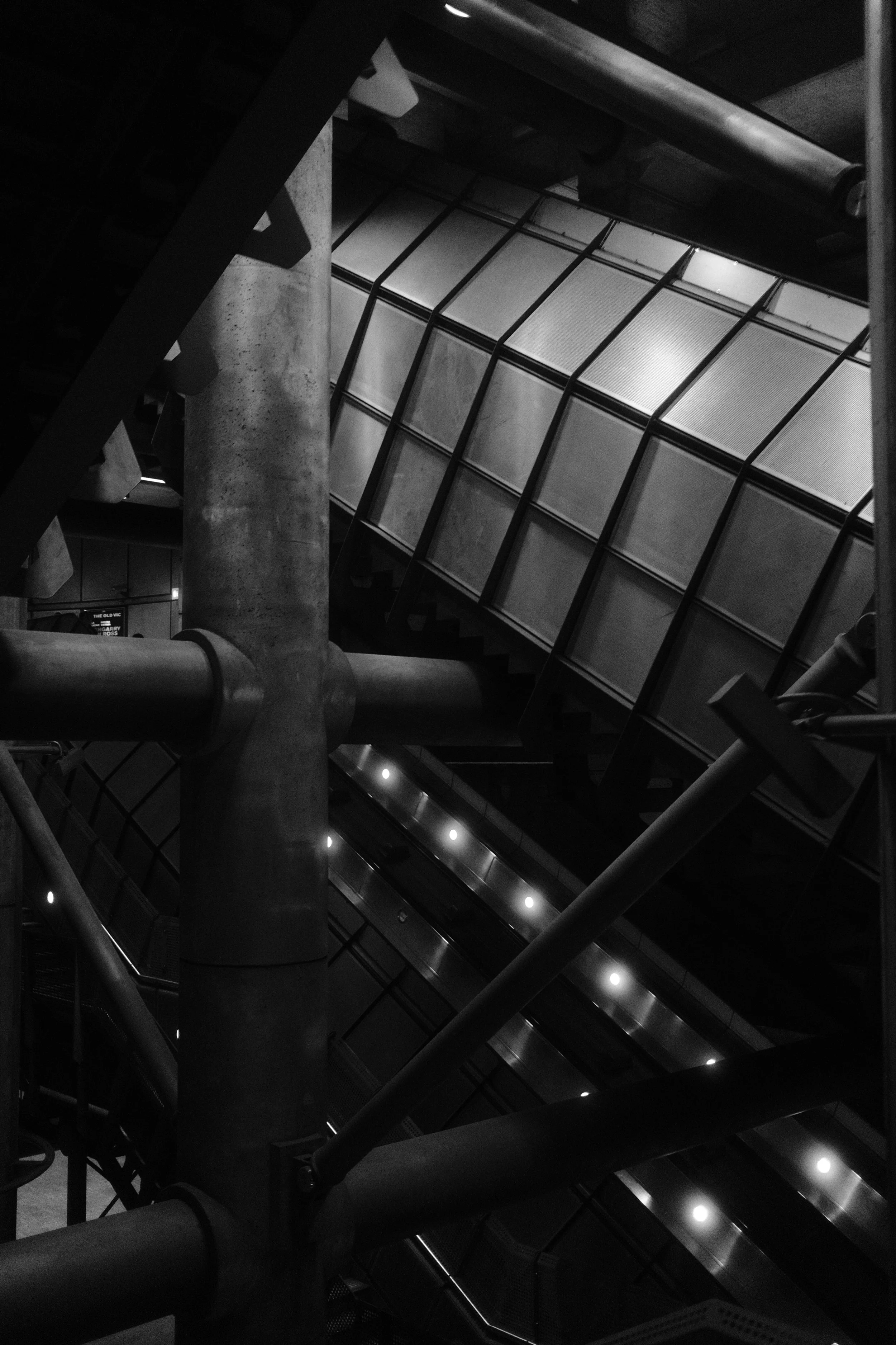 A black and white interior view of a modern building's escalators and structural elements, including large concrete columns, metal railings, and glass panels.