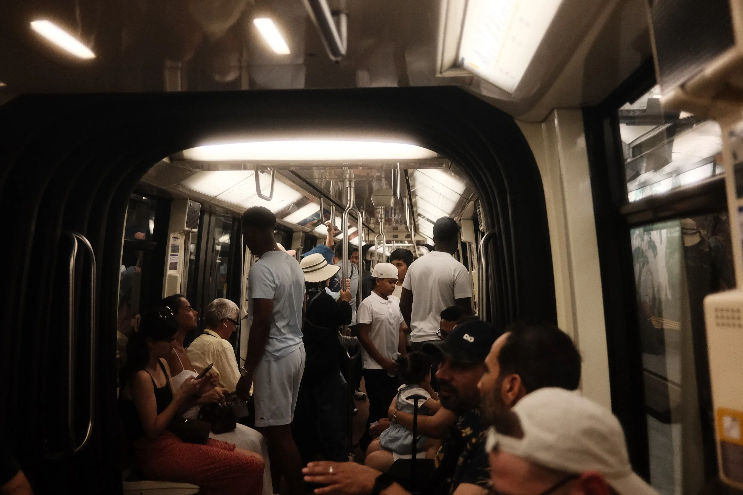 Crowded subway train with passengers standing and seated, people looking at phones, facing different directions, with overhead handles visible.