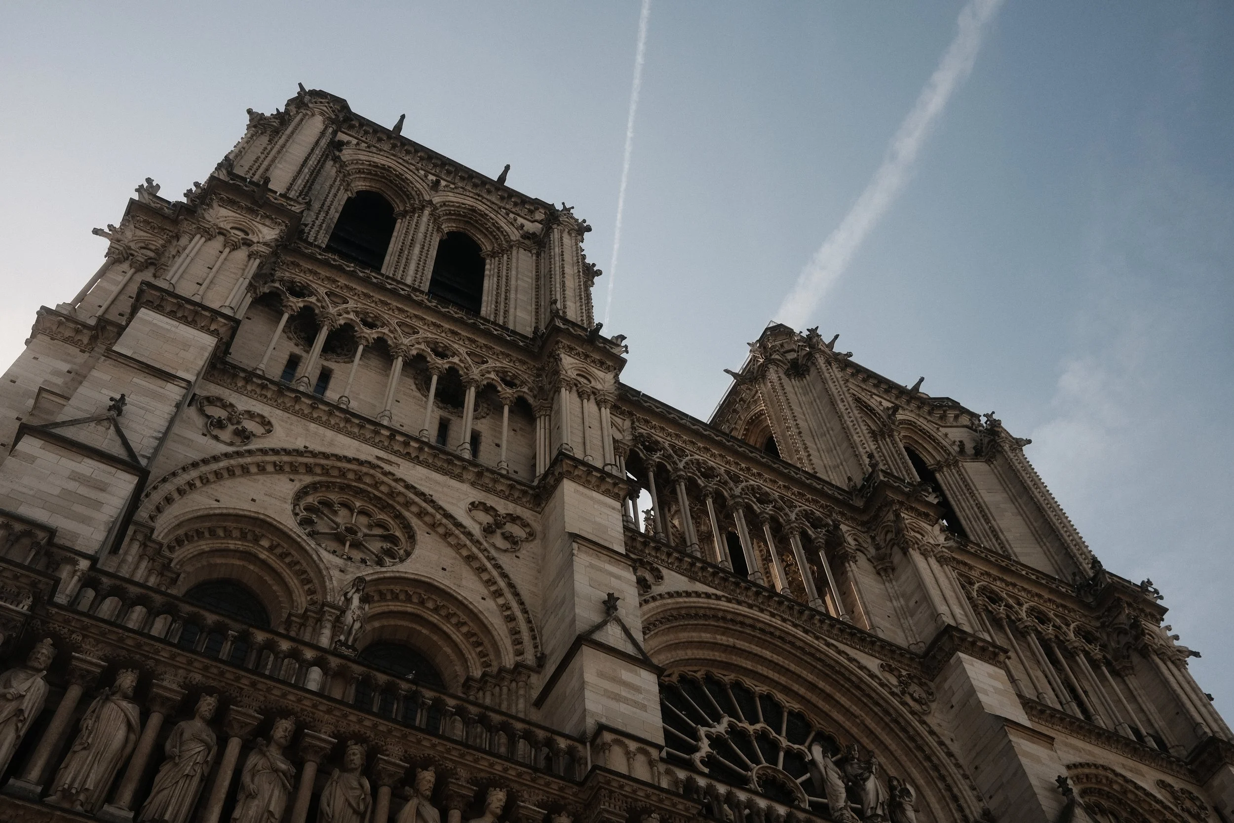 Close-up view of a historic Gothic-style cathedral with intricate stone carvings and statues on the facade, under a partly cloudy sky with visible contrails.