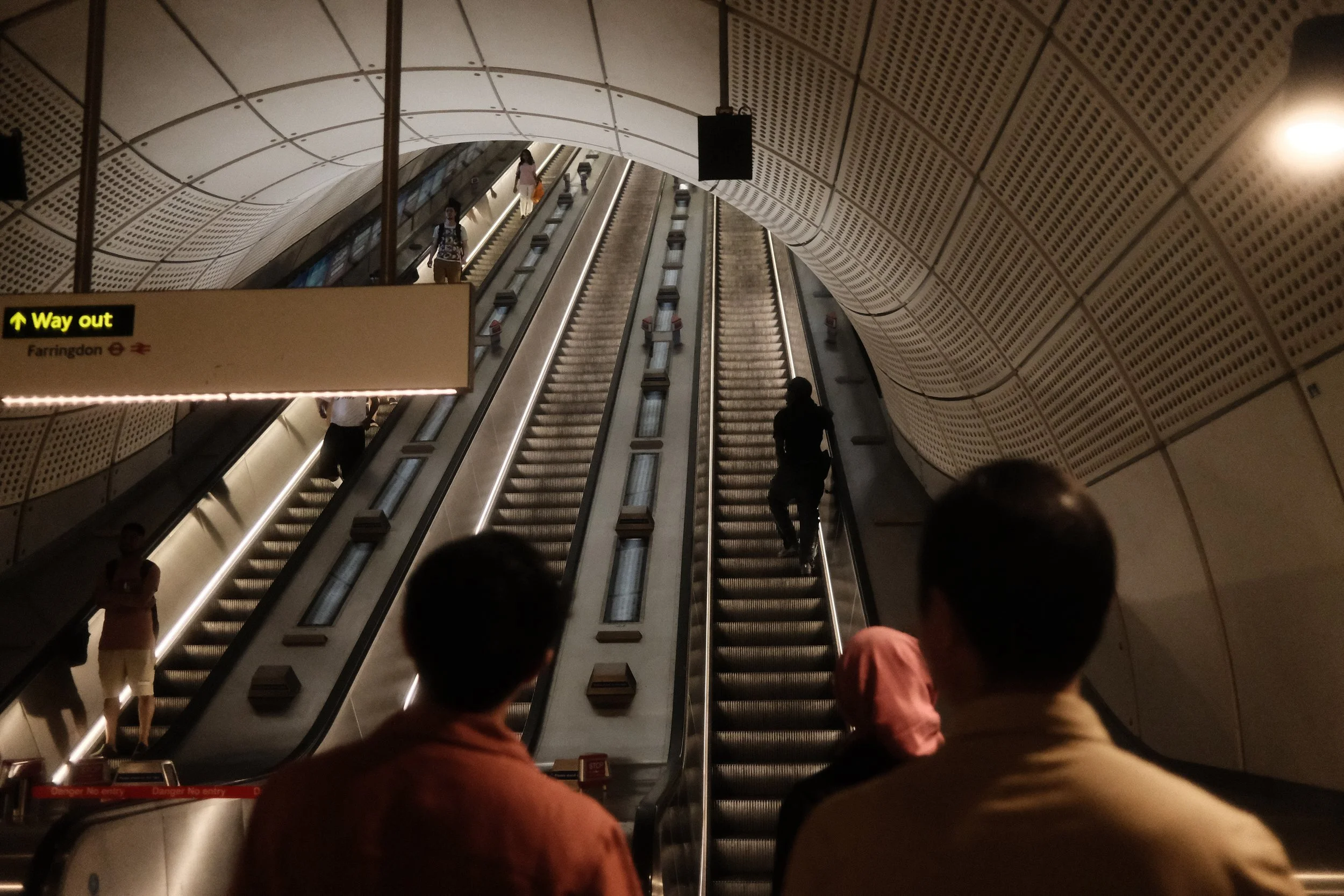 People riding escalators in a modern underground subway station, with a curved, perforated ceiling and a sign indicating the way out to Farringdon.