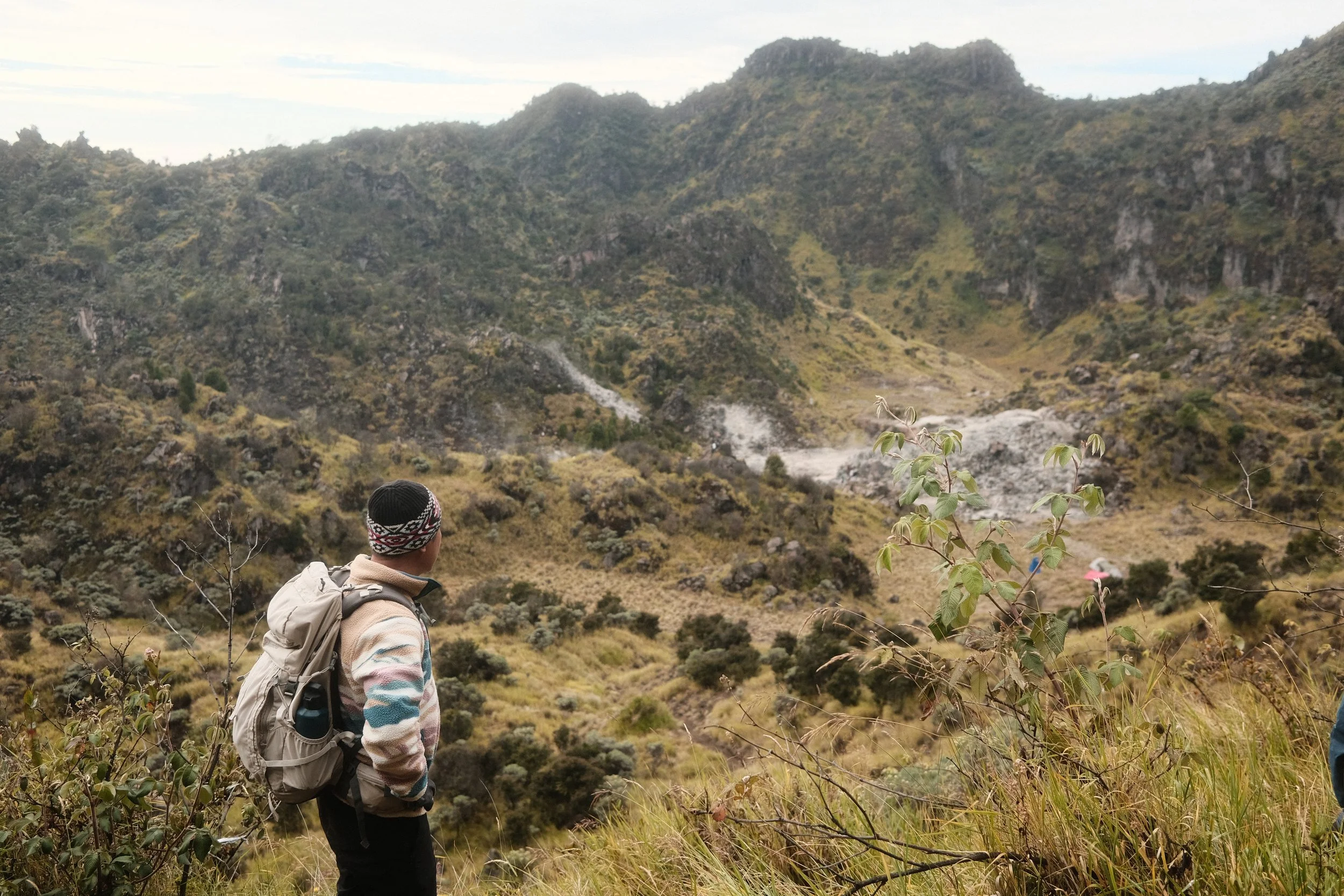 Hiker with a backpack and knit hat stands on a grassy trail overlooking a mountainous landscape with green vegetation and a cloudy sky.
