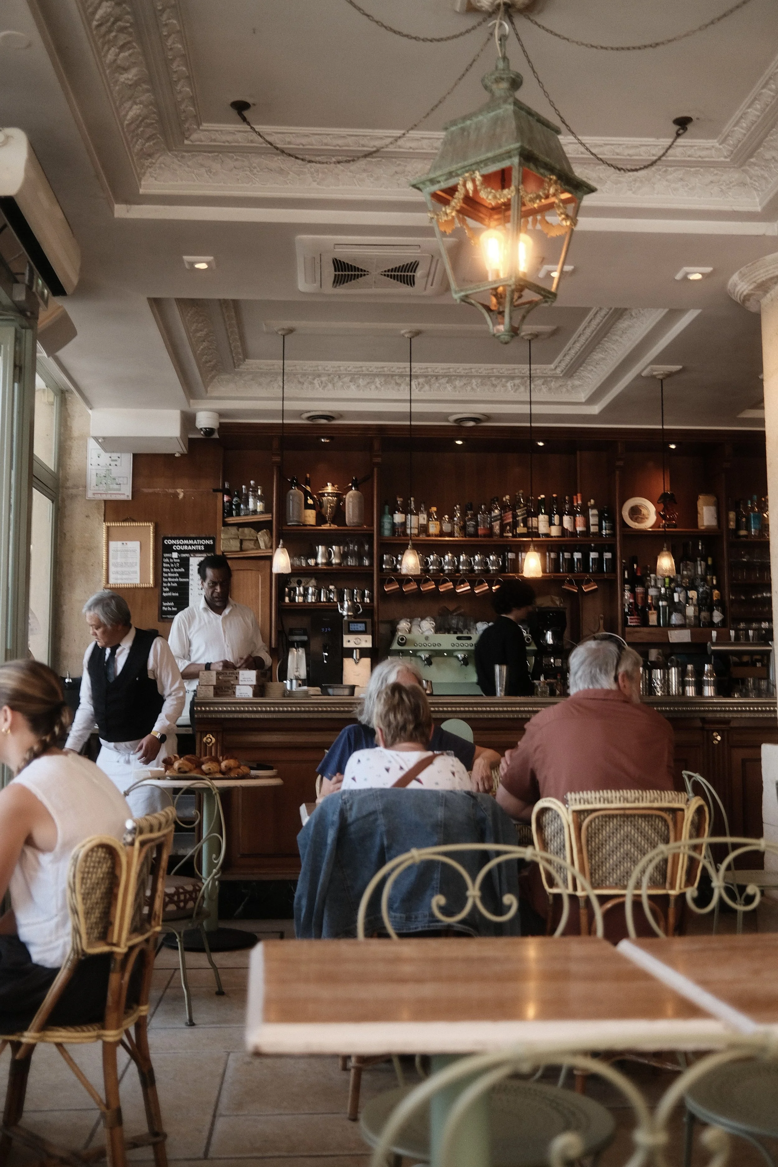 Inside a restaurant or café with ornate ceiling decor, hanging lamps, a bar area with bottles, and waitstaff attending to customers. Customers are seated at tables with wicker chairs, and a waiter is serving food.