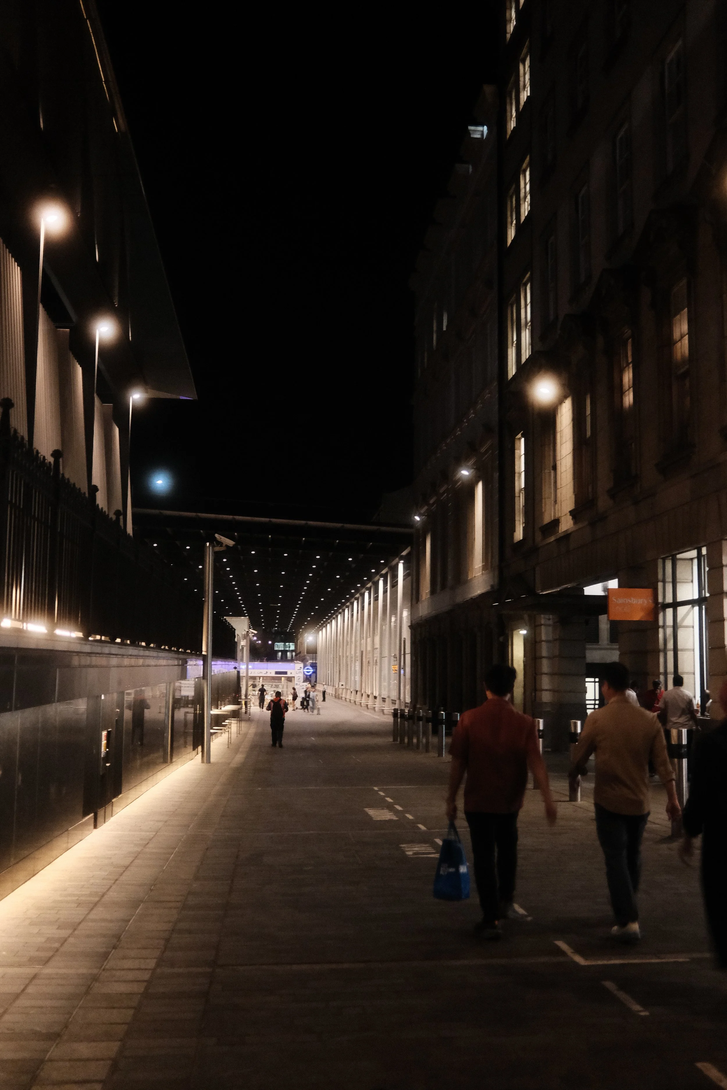 Nighttime scene of a city street with people walking on the sidewalk. The street is lit by streetlights and building lights, with tall buildings on either side. Some windows in the buildings are illuminated.