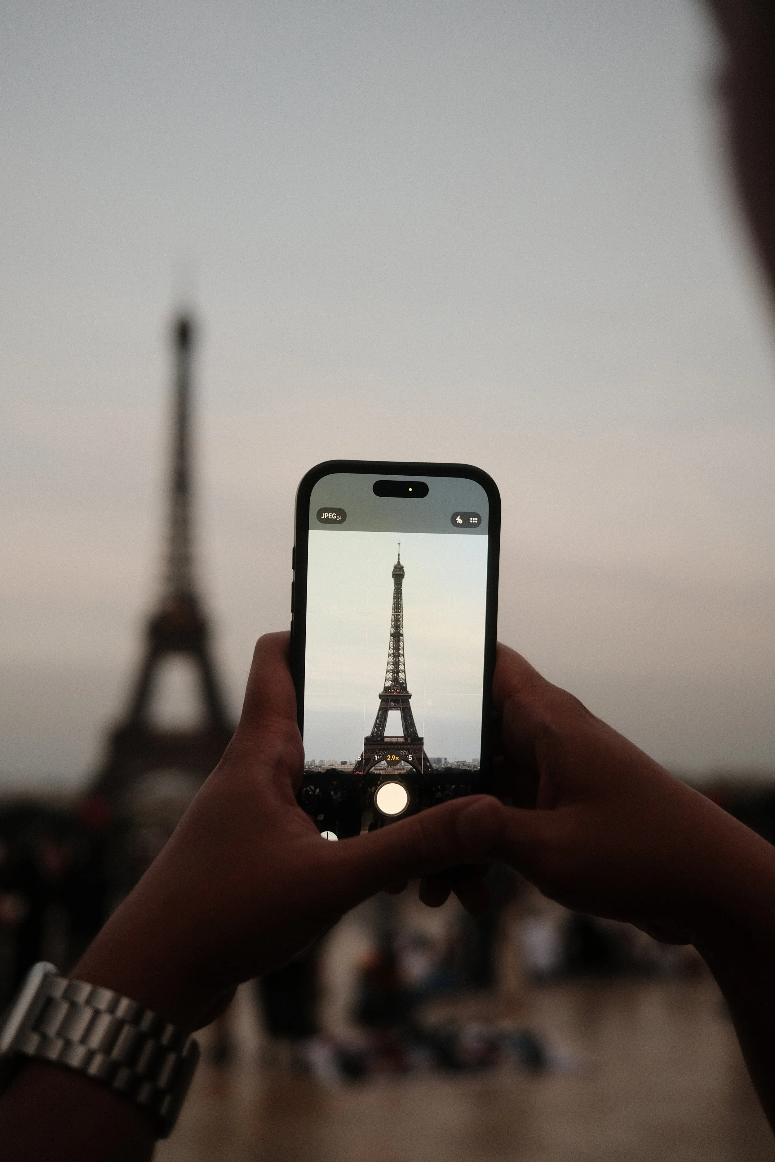 The image shows a person holding a smartphone taking a photo of the Eiffel Tower. The photo captures the tower framed on the phone's screen, with a blurred background and the tower centered, against a gray sky.
