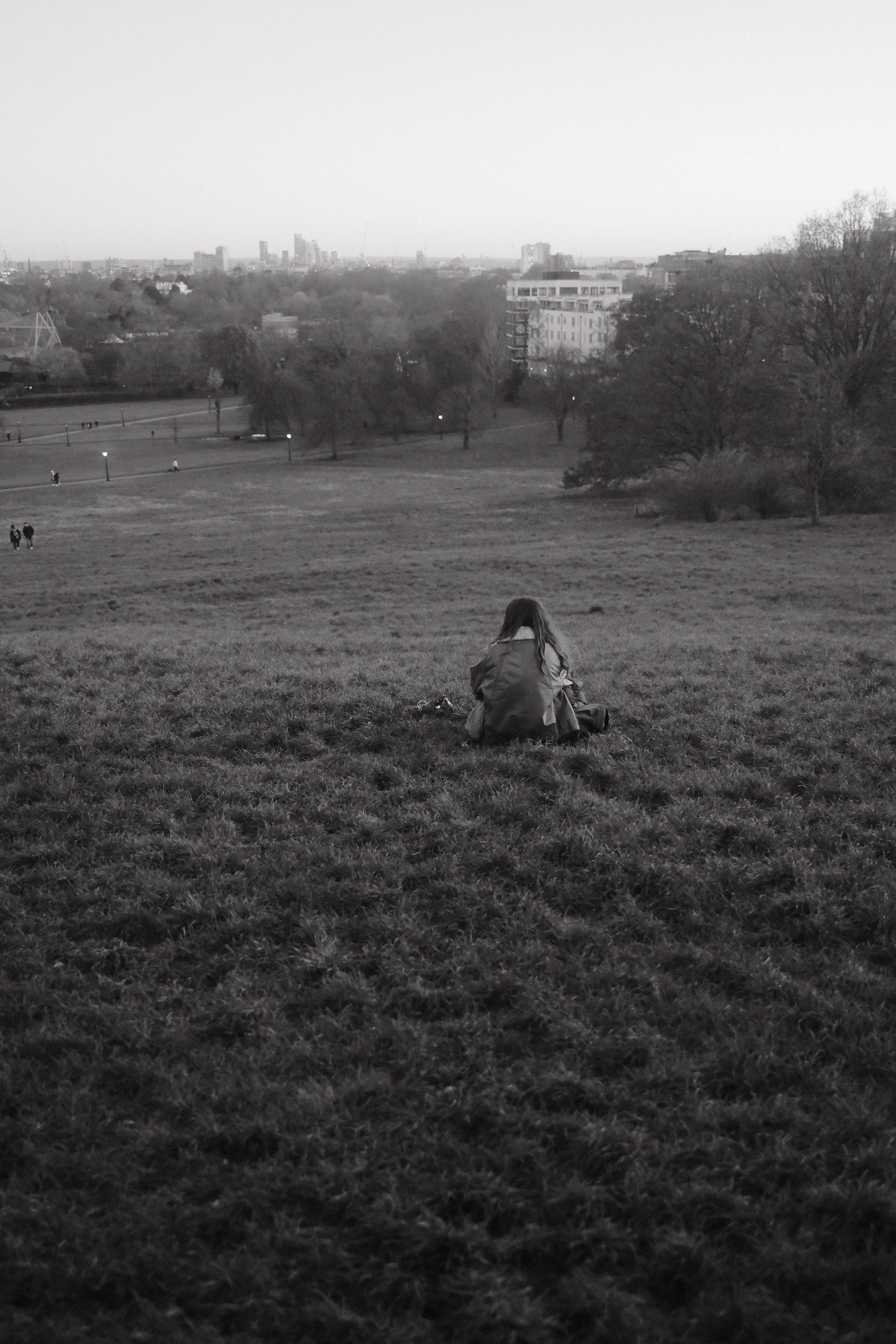 A person with long hair sitting on the grass in a park, with city buildings in the background, during the daytime.