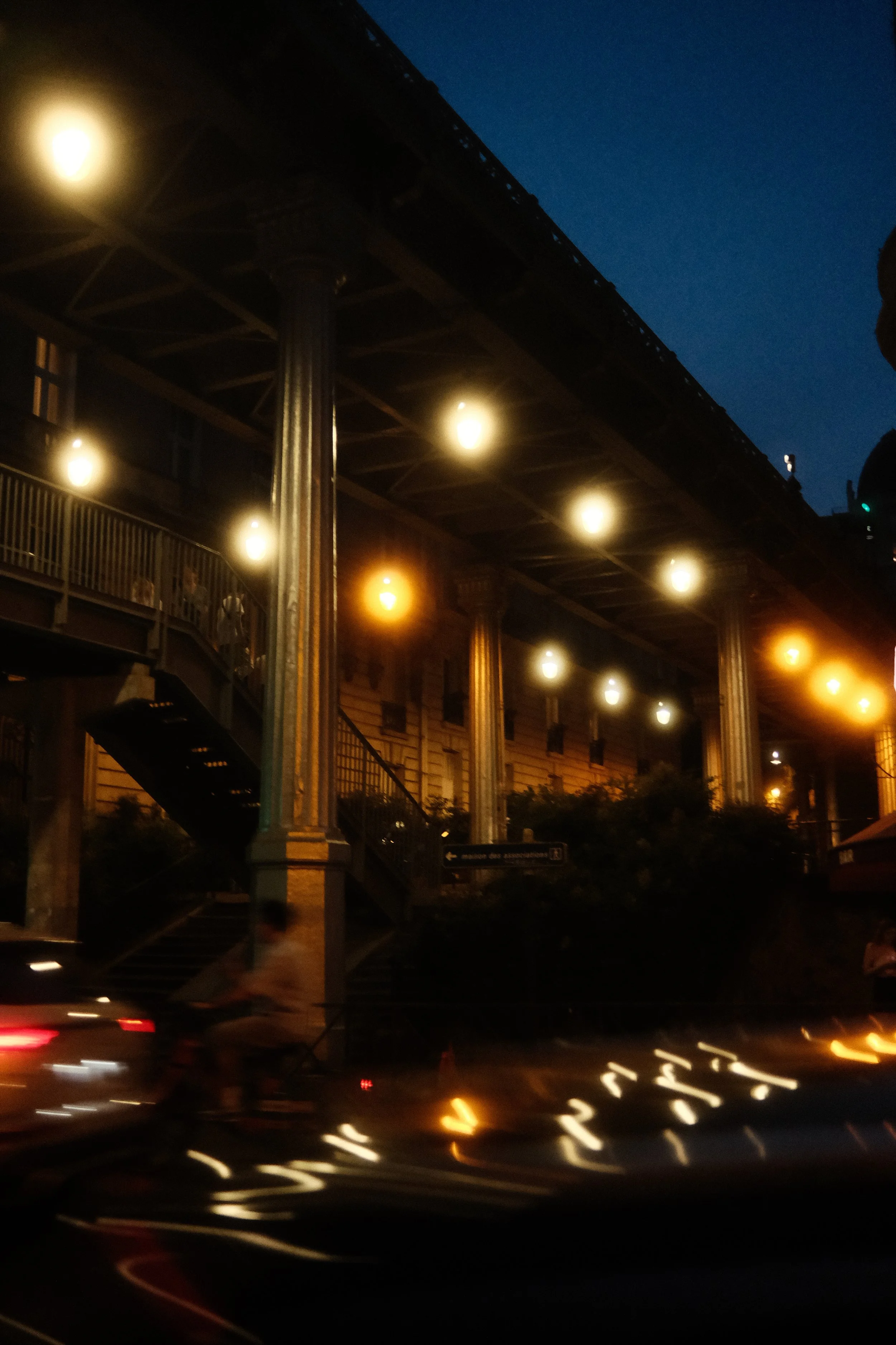 Nighttime city scene with illuminated streetlights, a building with columns and an overhead bridge, and blurry cars moving on the street.