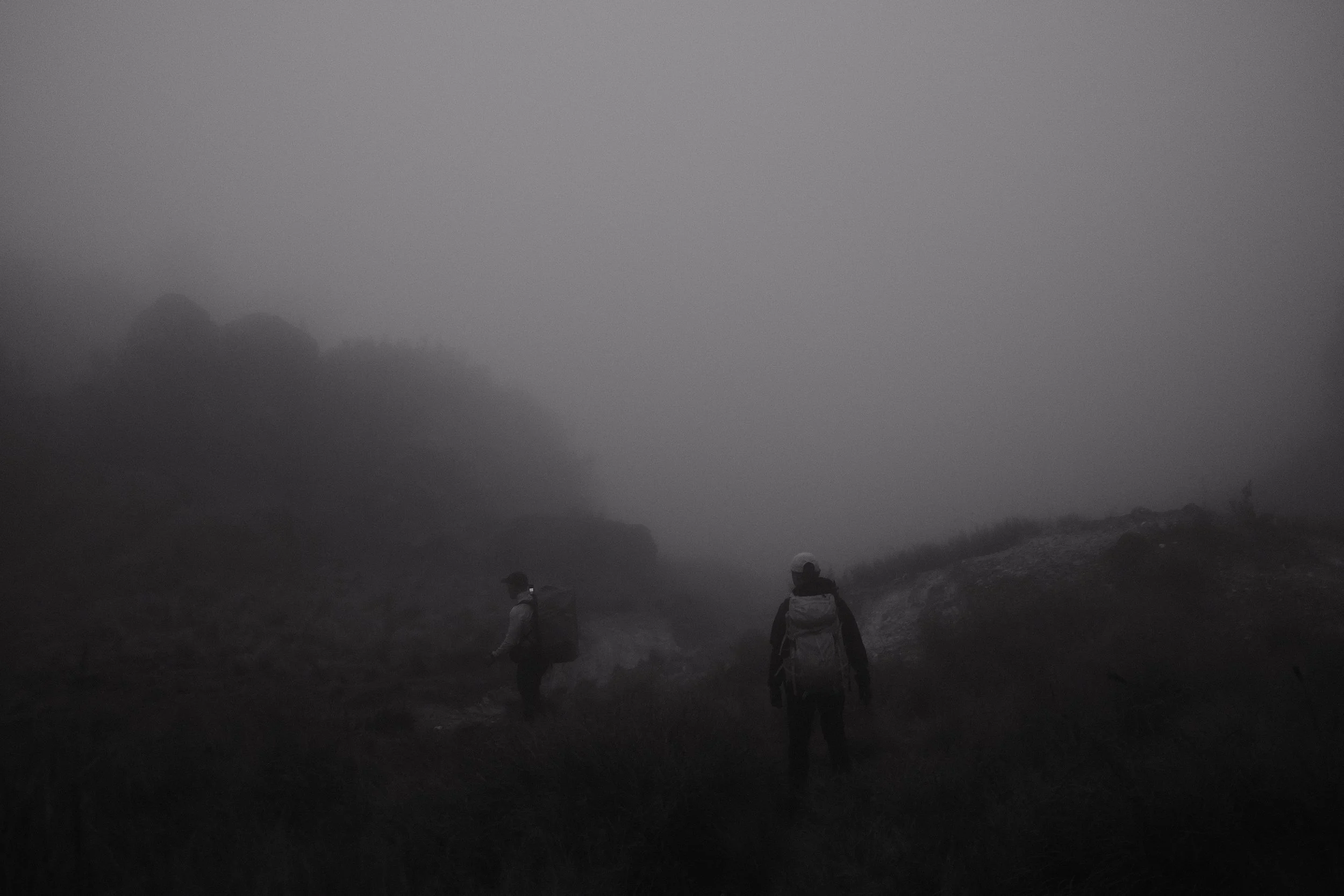 Three hikers walking on a foggy mountain trail, wearing backpacks and outdoor clothing, with fog-covered mountains in the background.