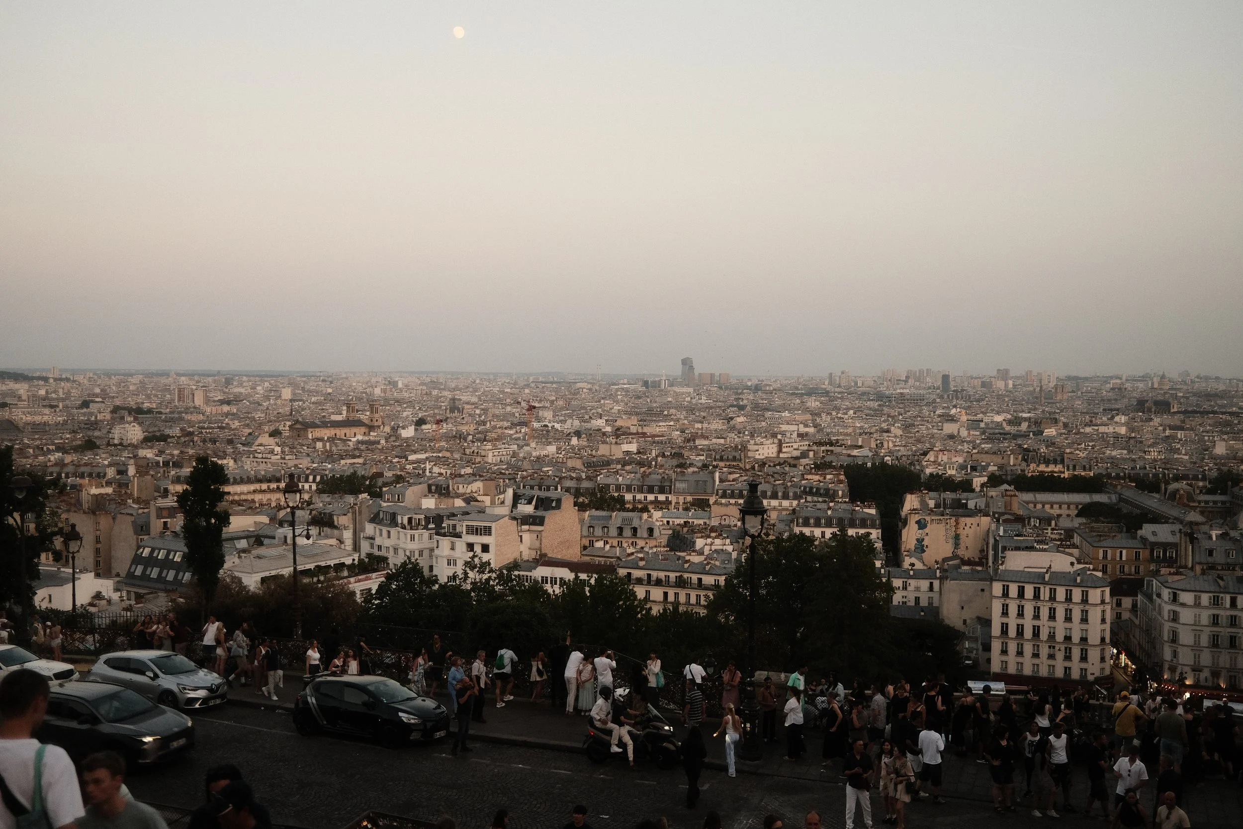 A panoramic view of Paris cityscape during sunset, with numerous buildings, streets, and the landmark La Défense business district visible in the distance. People are gathered at a viewpoint in the foreground, and cars are parked along the street.