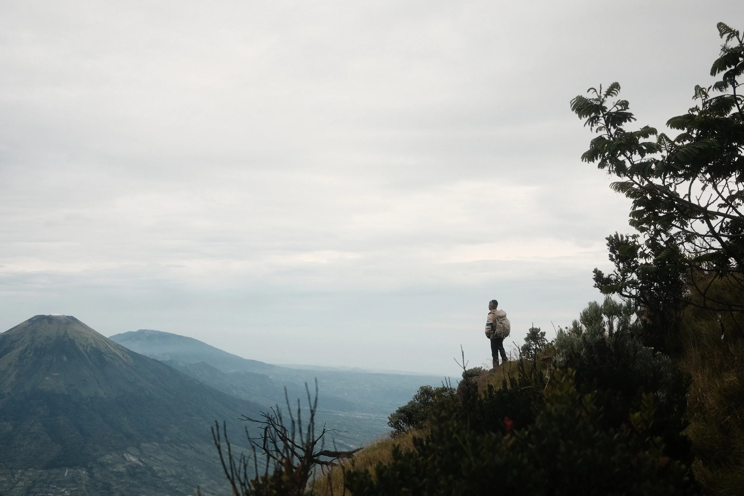 A man standing on a trail overlooking mountains and valleys under a cloudy sky, wearing a backpack and outdoor clothing.