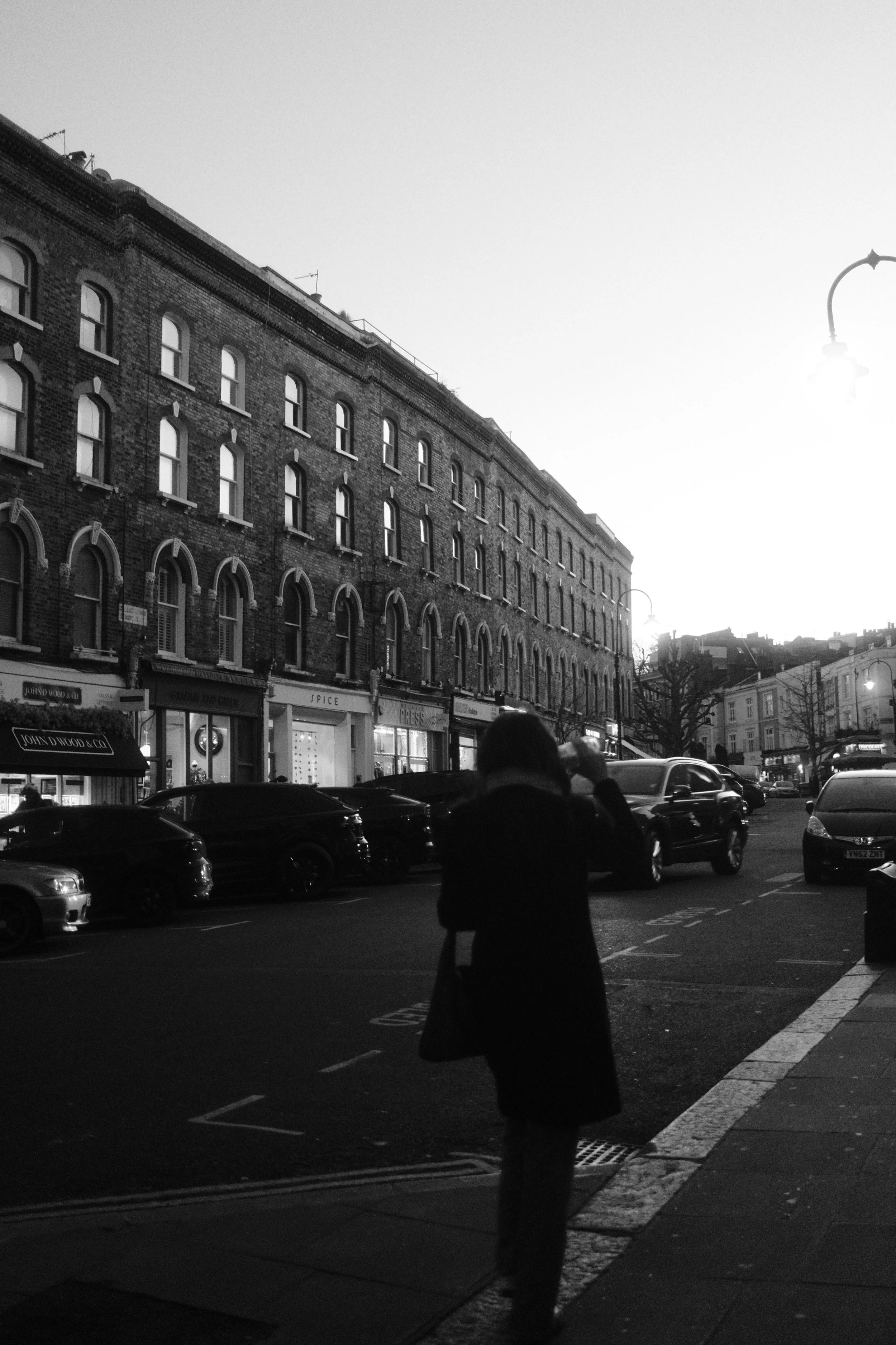 Black and white photo of a city street with older brick buildings, cars parked along the street, and a woman in the foreground holding a coffee cup, with a bright sky overhead.
