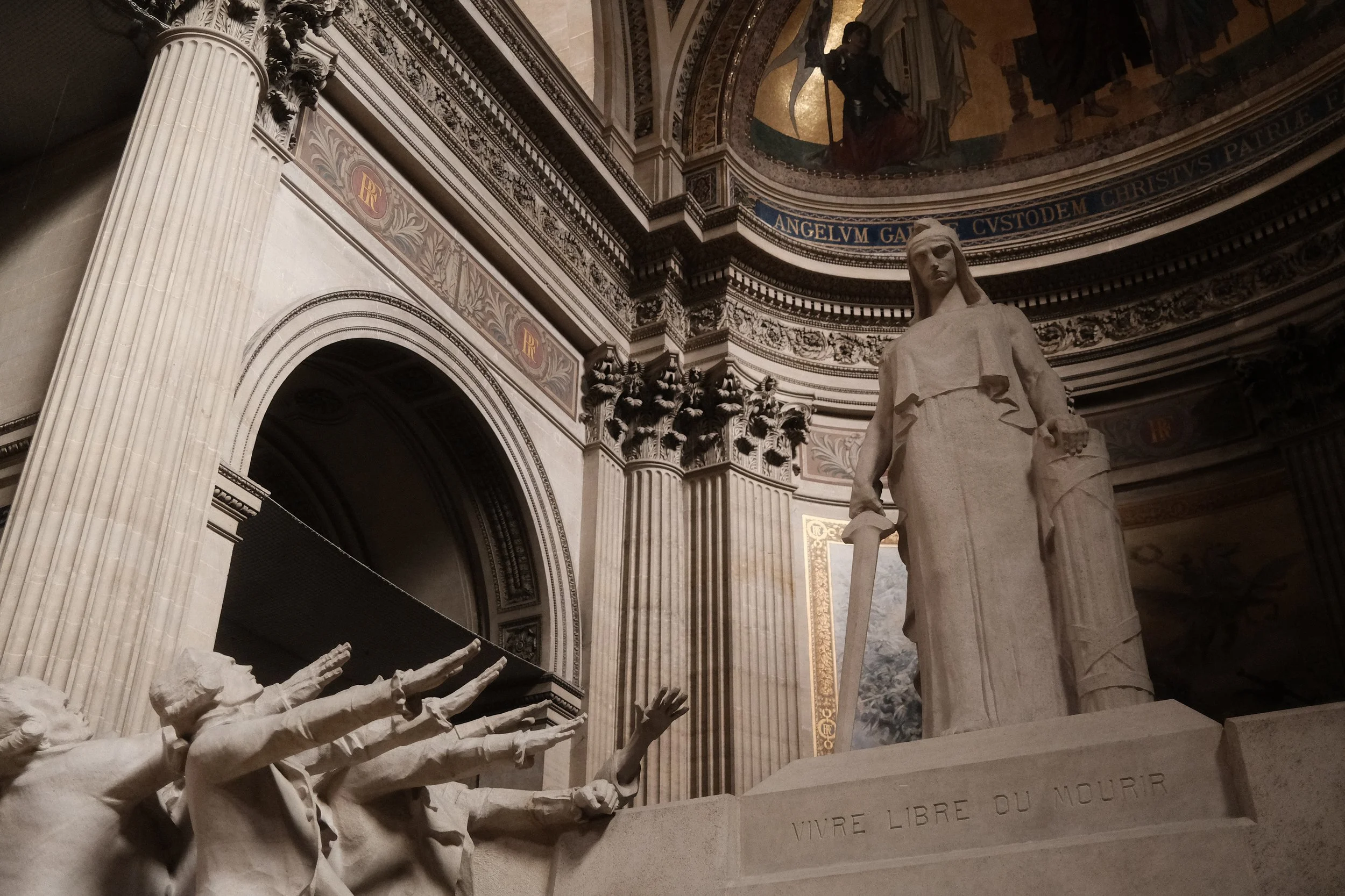 Interior of a grand classical building featuring a white marble statue of a woman holding a sword, surrounded by ornate architectural details and Latin inscriptions, with a domed ceiling and a painting or mural at the top.