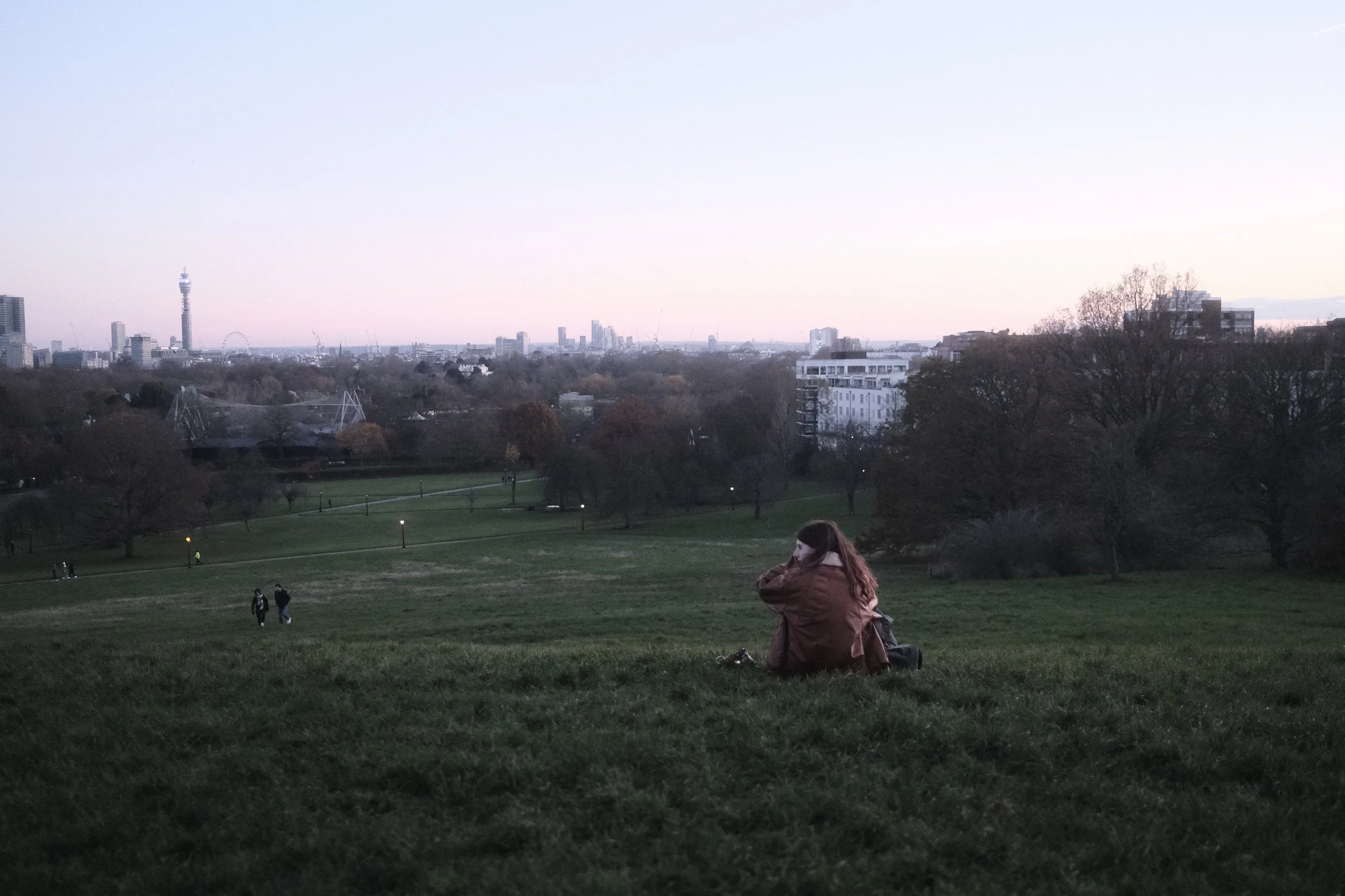 A woman with red hair sitting on the grass in a park, looking towards the city skyline in the distance during dusk, with a few other people also enjoying the park.
