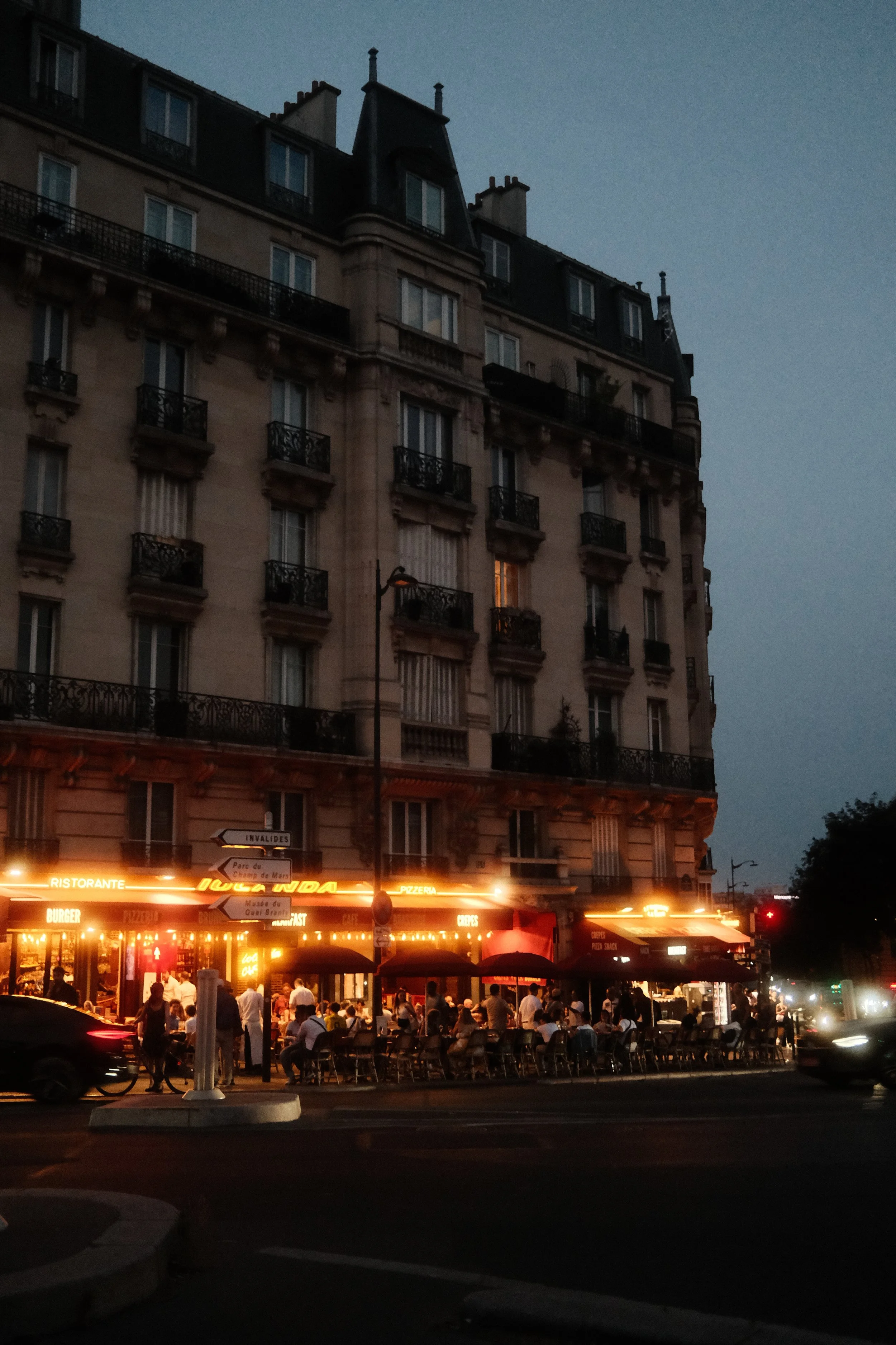City street scene at dusk with a tall, classic European style building and a busy outdoor restaurant with bright neon signs and outdoor seating, and people walking and dining.