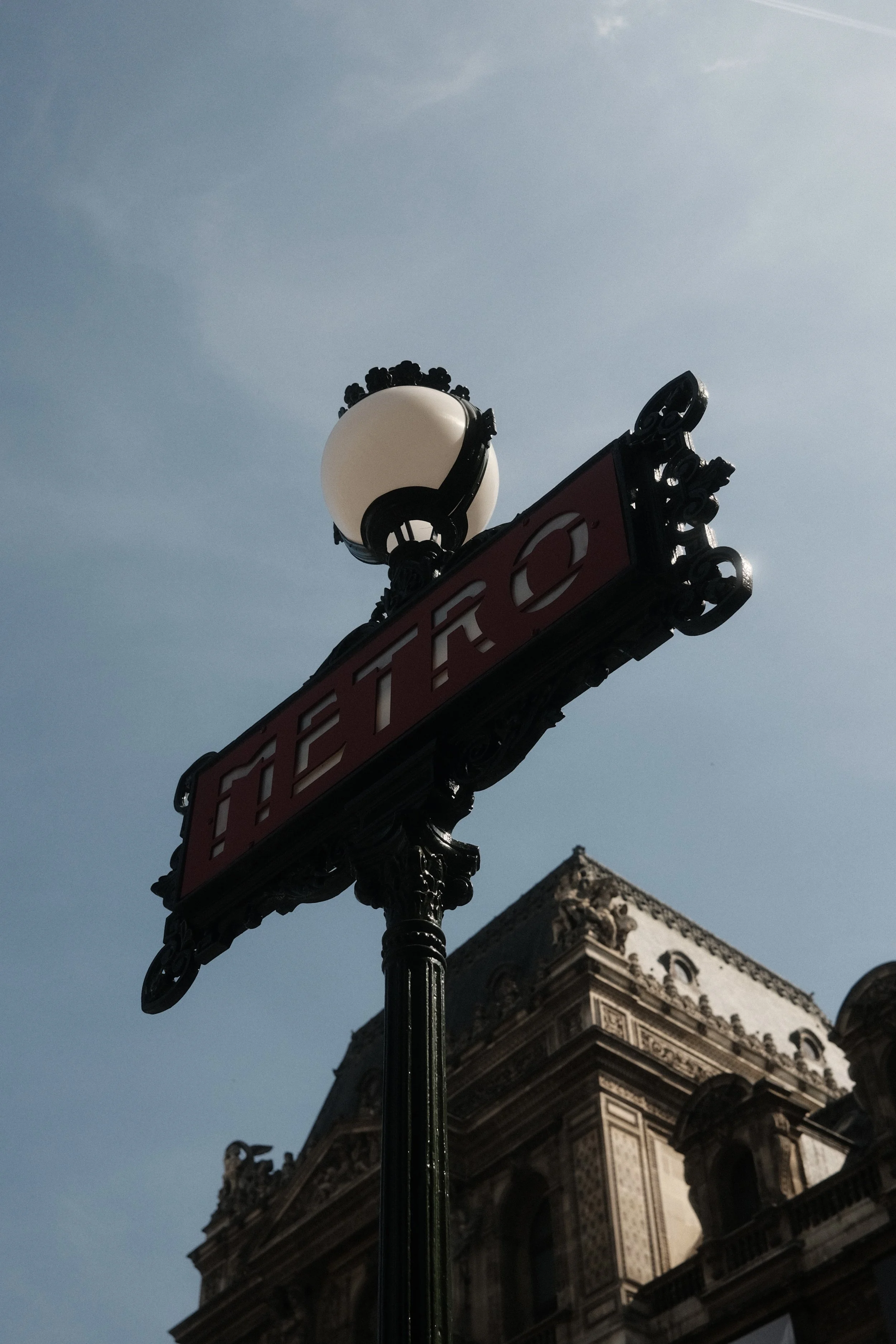 Street sign with red background and white text reading 'THE'. An ornate street lamp with a round white globe is mounted above the sign. In the background, part of a historic stone building with detailed architectural features is visible, set against 